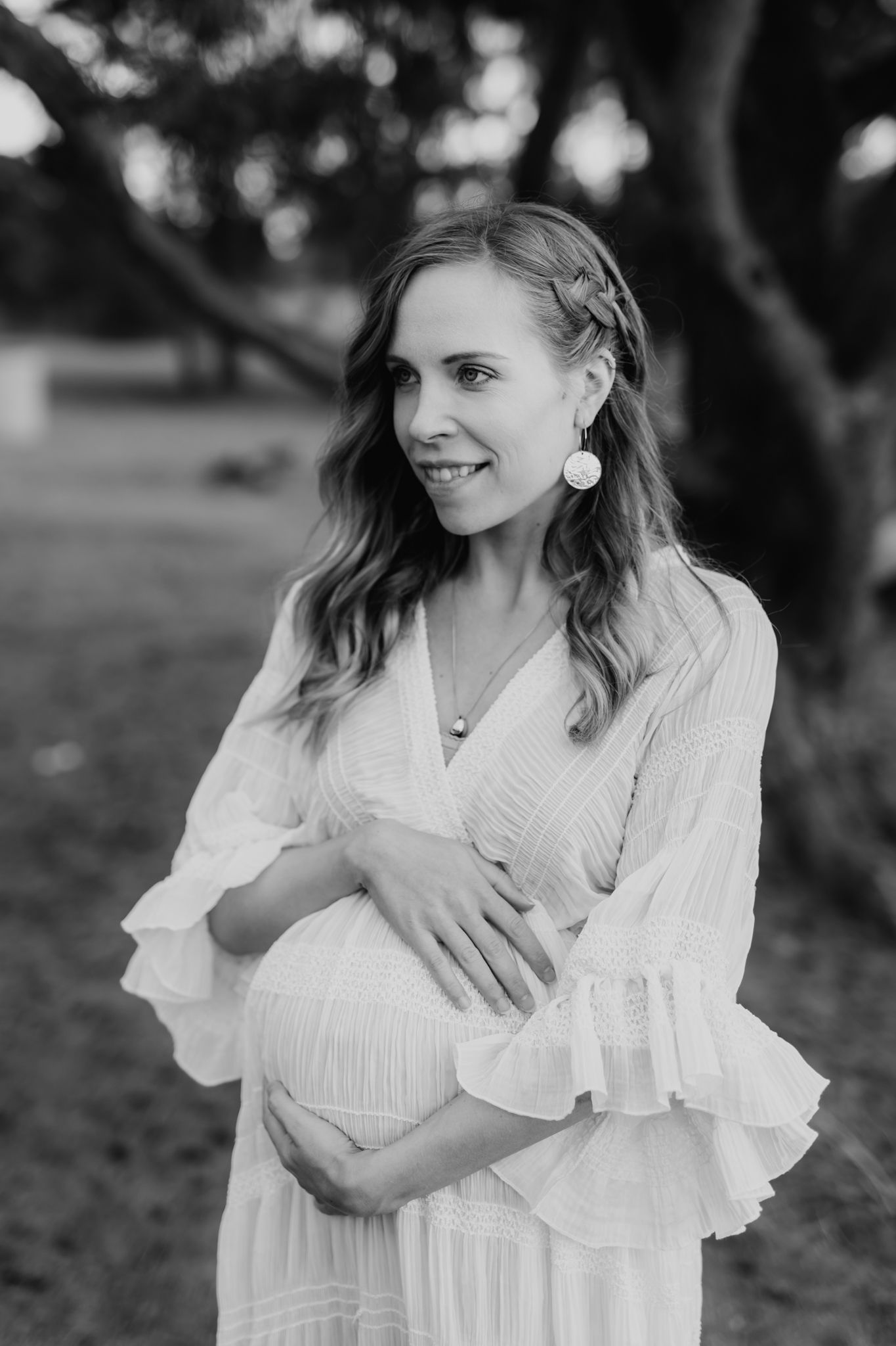 Smiling pregnant woman in white dress holding baby bump outdoors in black and white maternity photo.