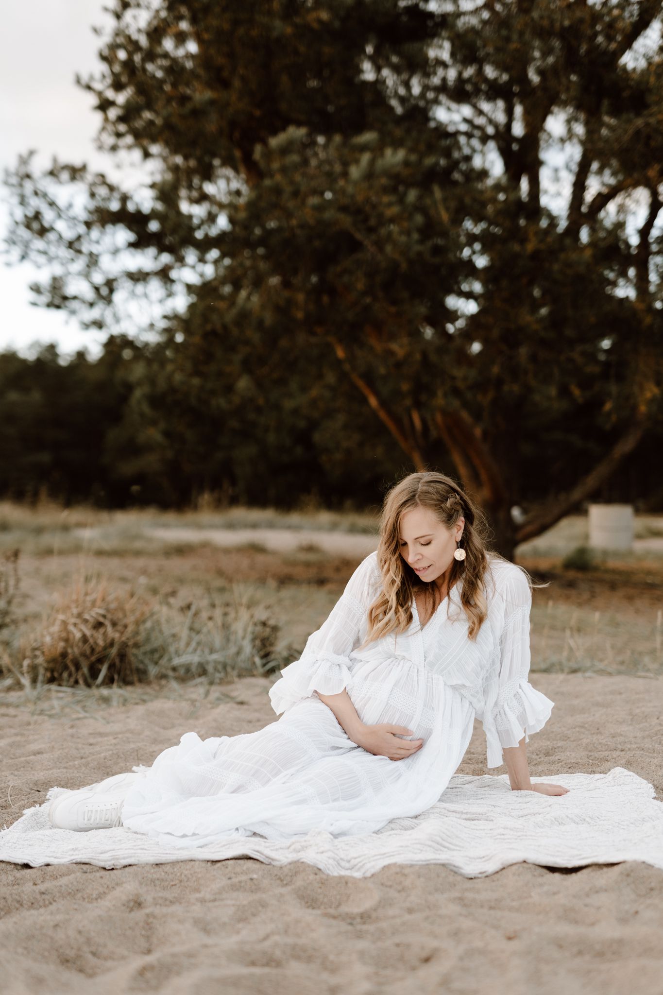Pregnant woman in flowing white dress sitting on sandy ground outdoors near large trees.