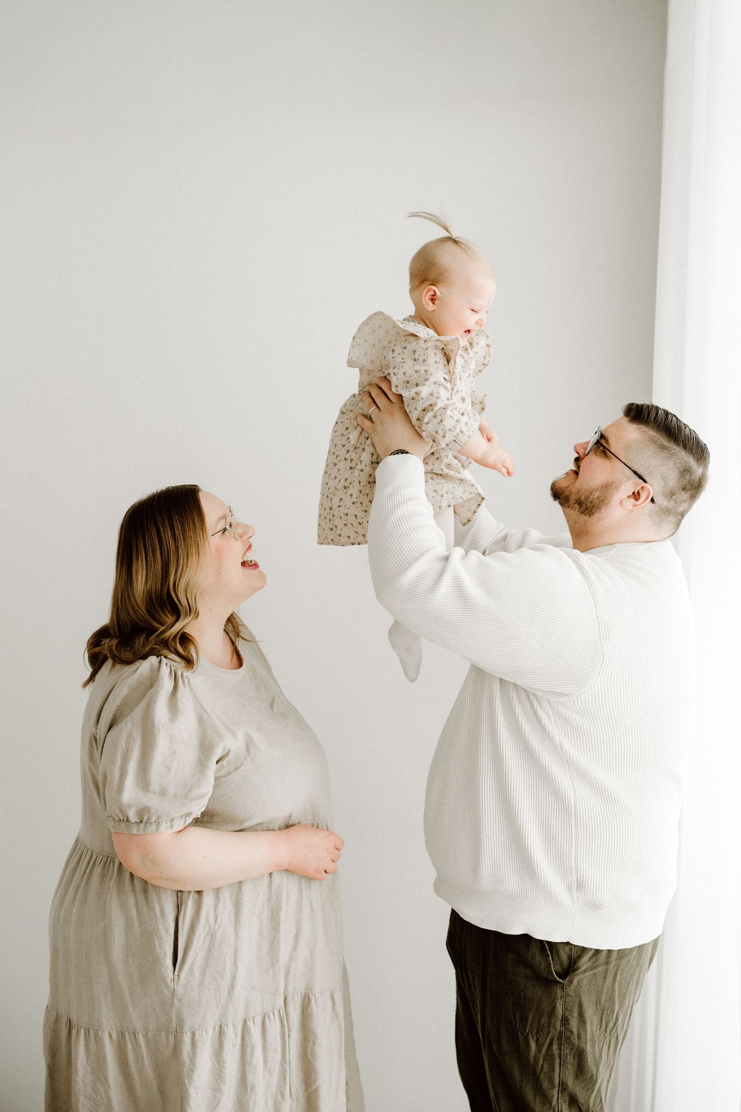 A pregnant woman in a linen dress watches as a bearded man in glasses lifts a smiling baby girl in a floral romper.