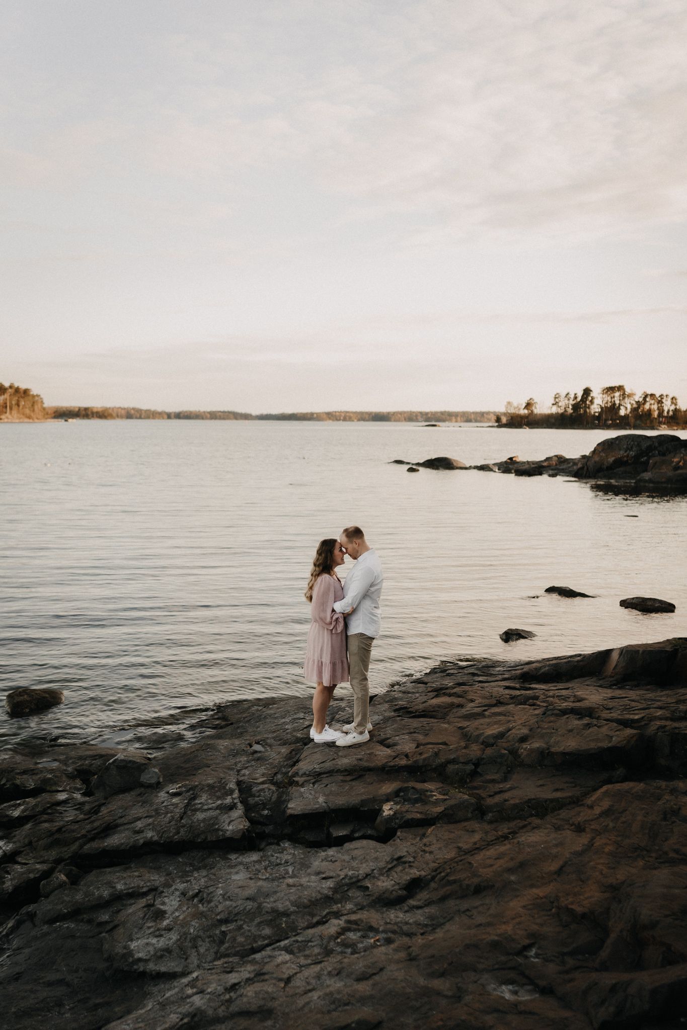 Couple embracing on rocky shoreline at sunset with calm lake and tree-lined horizon in background.