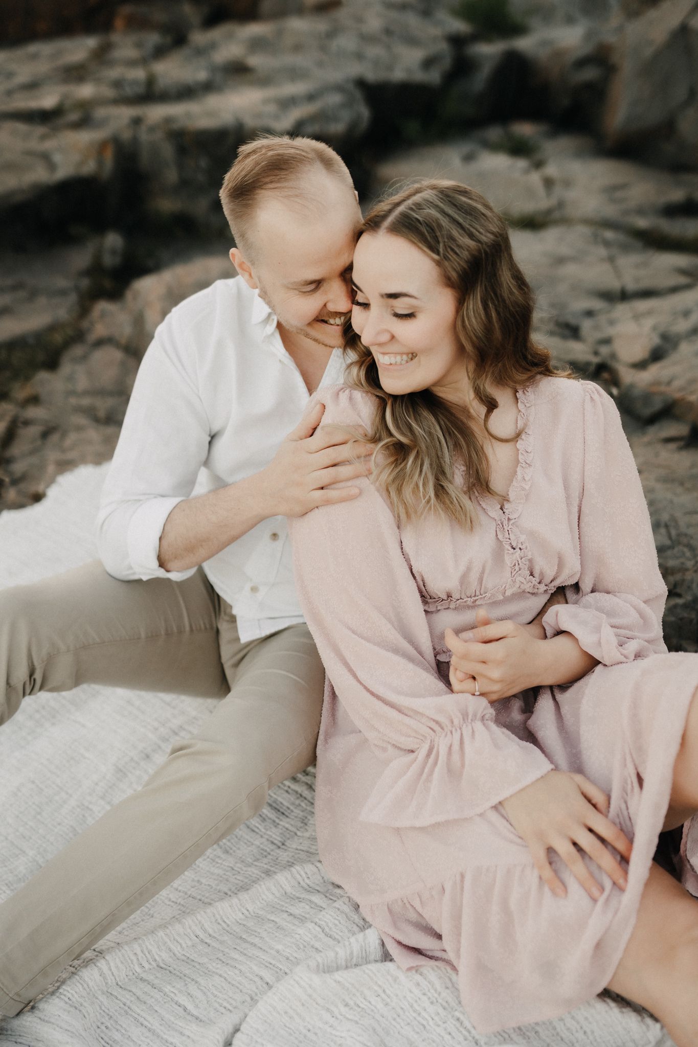 Couple cuddling on a white blanket by rocky shore, woman in pink dress smiling softly at sunset.