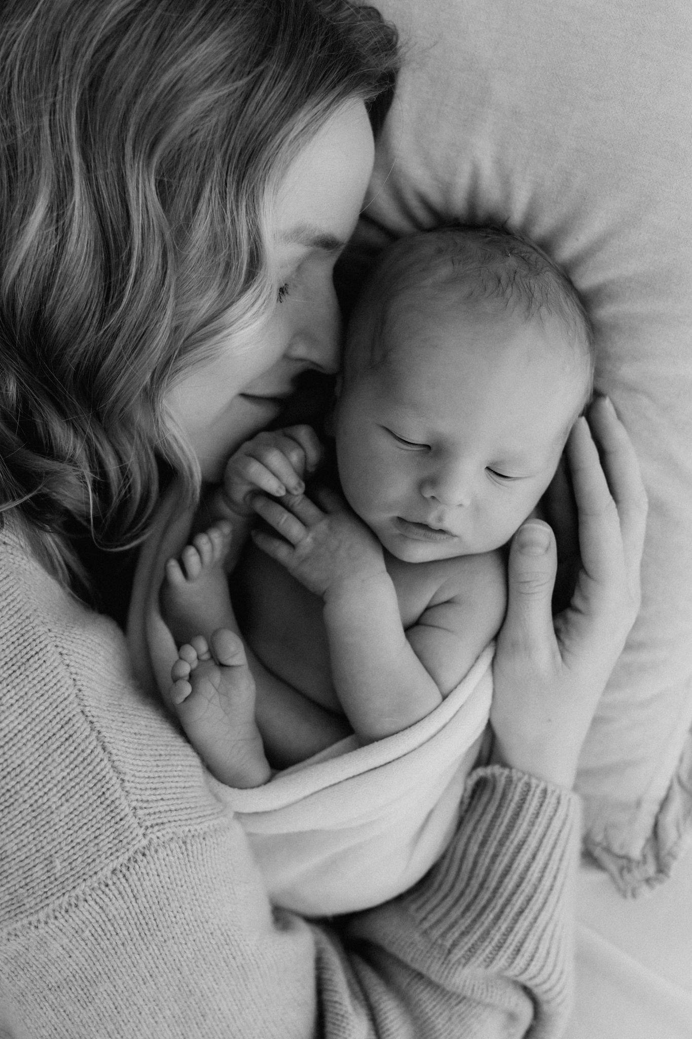 Black and white overhead photo of a mother nuzzling her sleeping newborn baby on a soft pillow.
