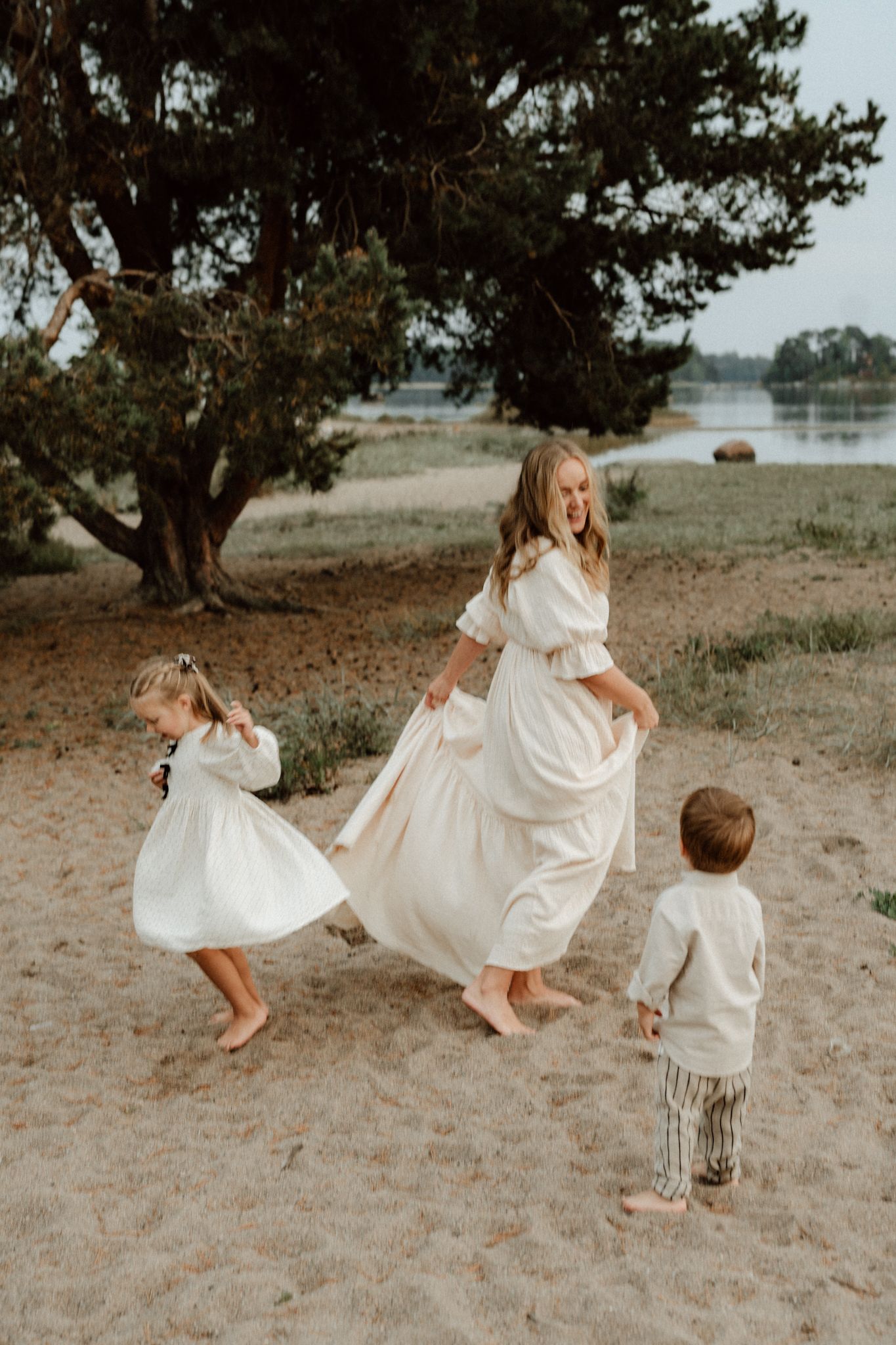 A smiling mother in a cream dress dances barefoot on sandy beach with her two young children.