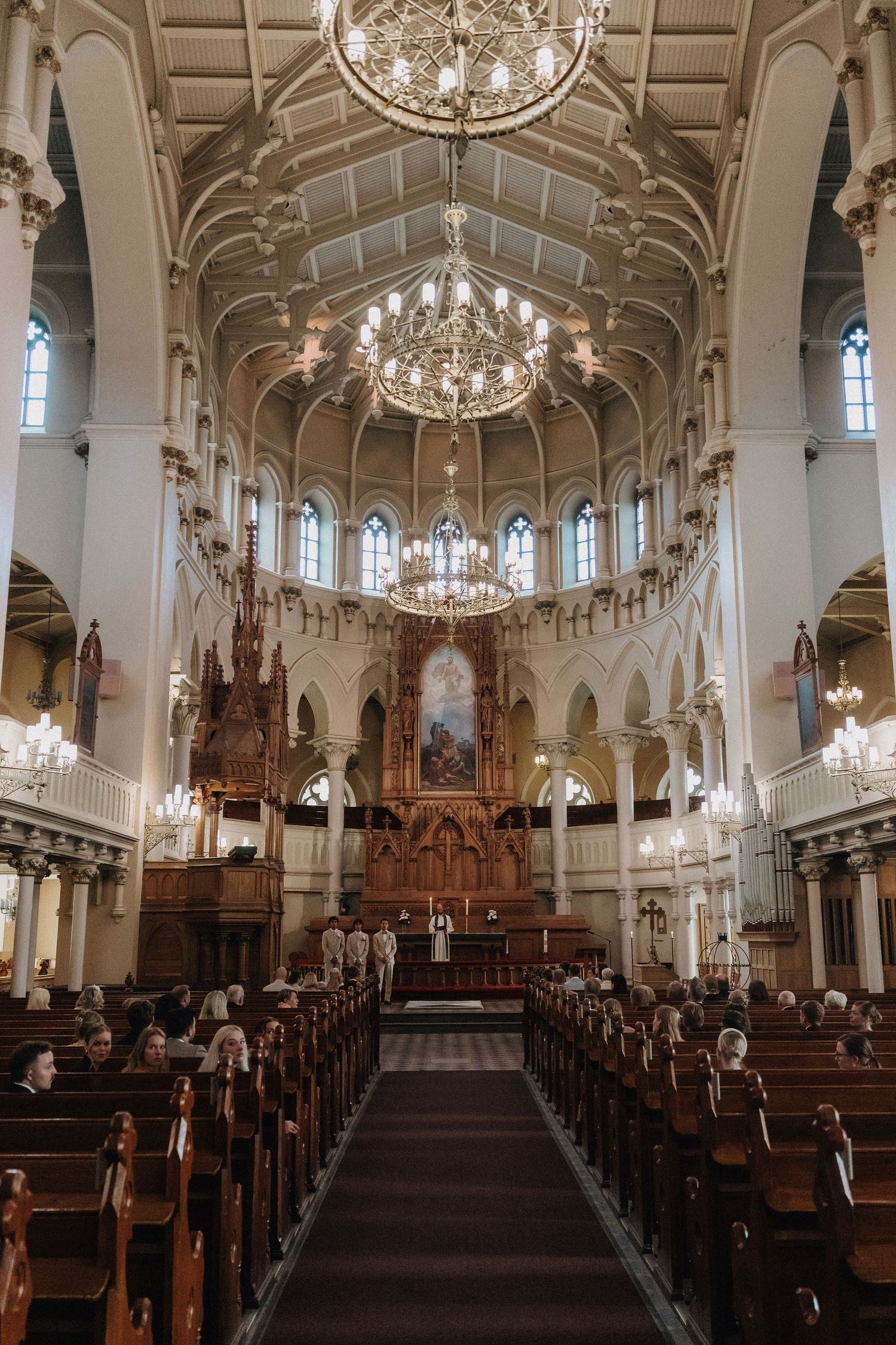 Elegant church interior with ornate wooden altar, crystal chandeliers, and rows of pews during a wedding ceremony.