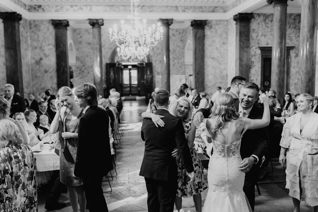 Black and white photo of newlyweds embracing guests in elegant marble wedding reception hall.