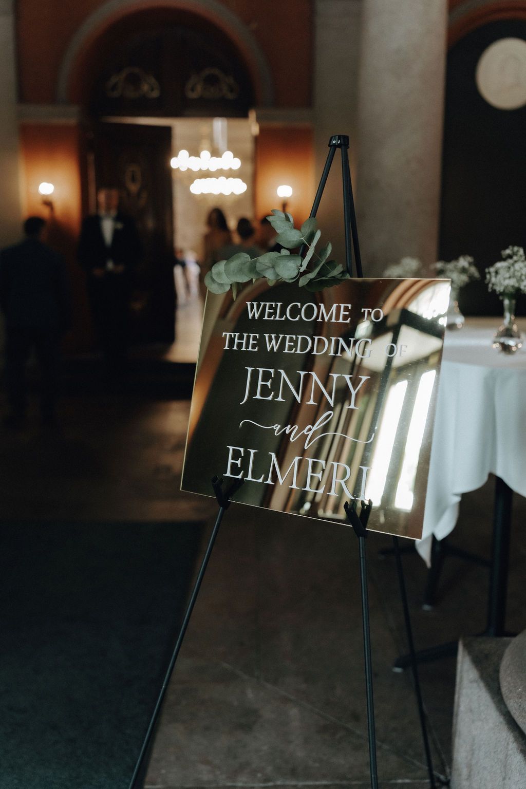 Elegant mirrored wedding welcome sign for Jenny and Elmeri on black easel with eucalyptus greenery.