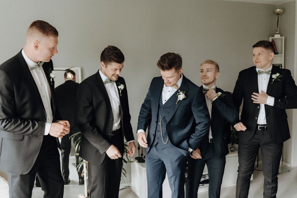 Five groomsmen in black and navy suits laugh together during wedding preparations.