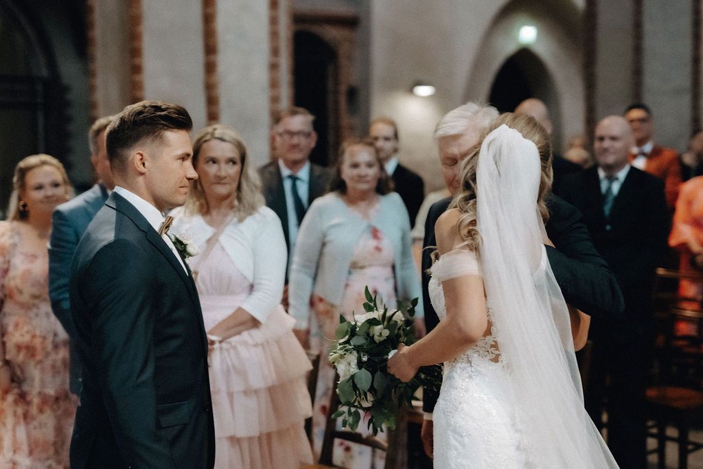 Groom in dark suit meets bride holding bouquet at church wedding ceremony aisle.
