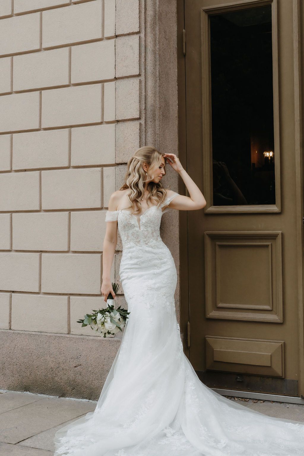 Elegant bride in off-shoulder lace mermaid gown holds white bouquet beside ornate building doorway.