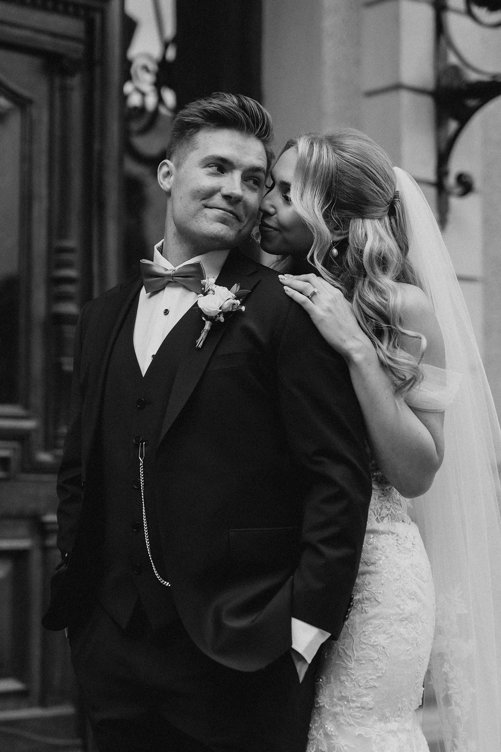Black and white close-up of smiling groom in tuxedo as bride in lace dress whispers in his ear.