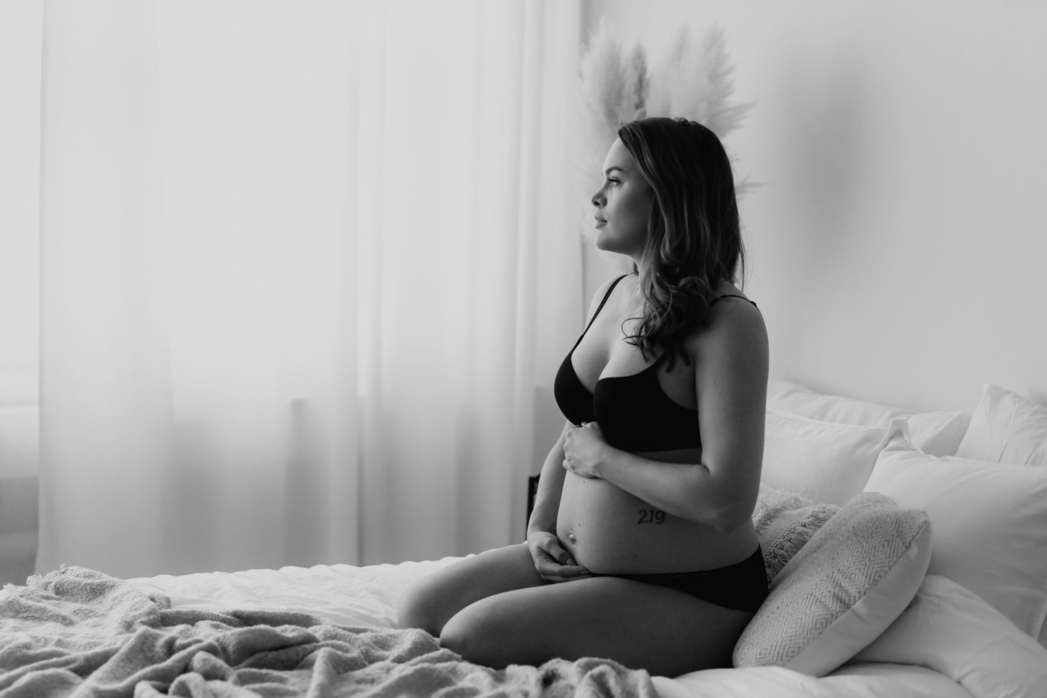 Black and white maternity photo of pregnant woman sitting on bed gazing out window.