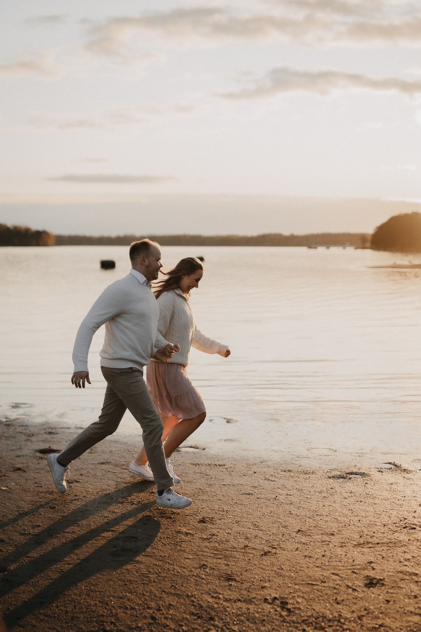 Couple runs playfully together along a sandy beach at sunset with warm golden light reflecting on water.