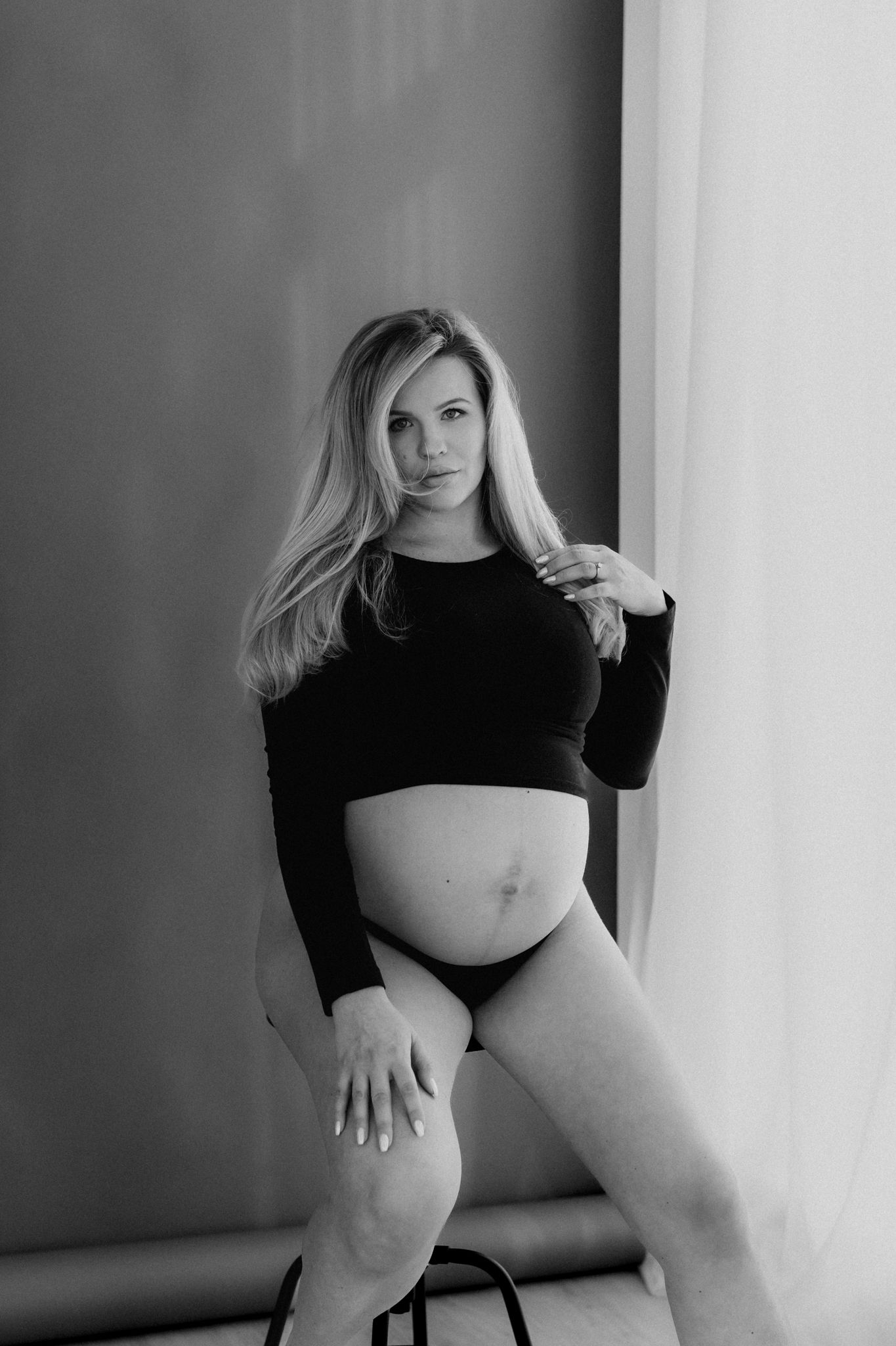 Black and white photo of pregnant blonde woman in black crop top seated on stool.