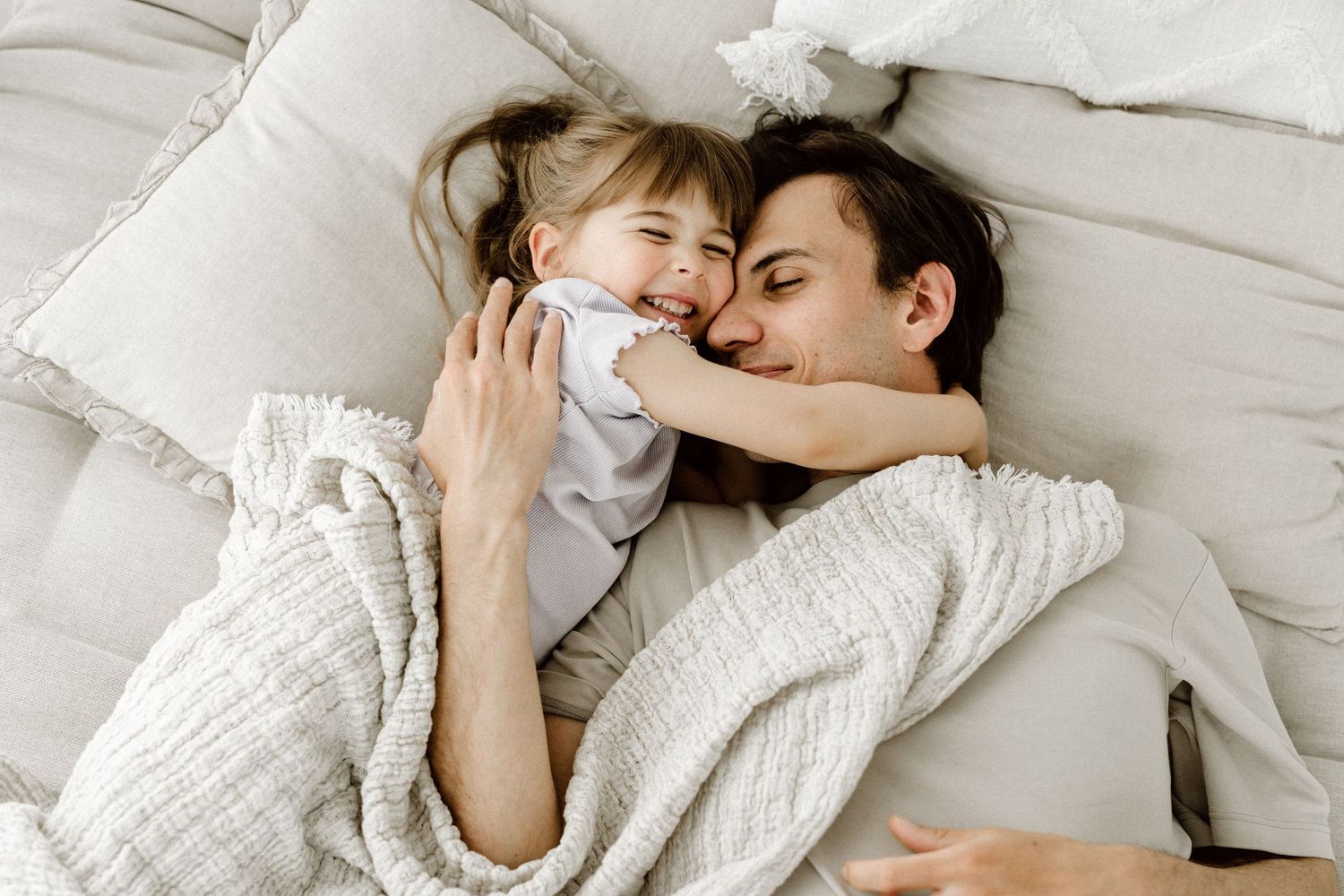 Mother and young daughter cuddling together smiling under cozy white blanket in bed.