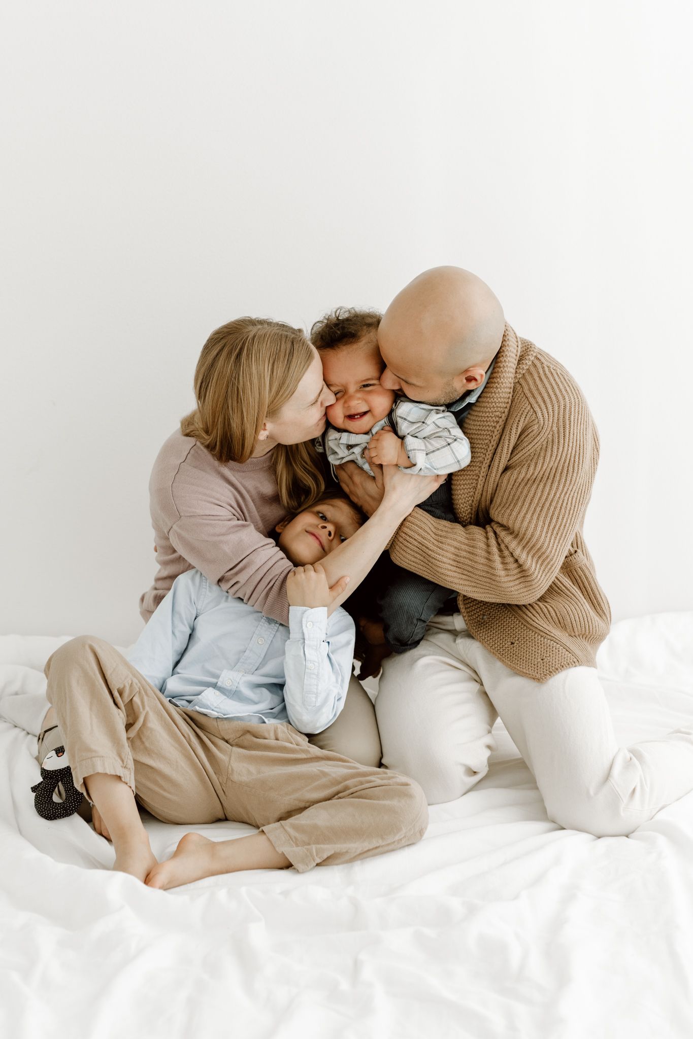 Loving parents kissing and cuddling their giggling child on a white bed in a bright room.