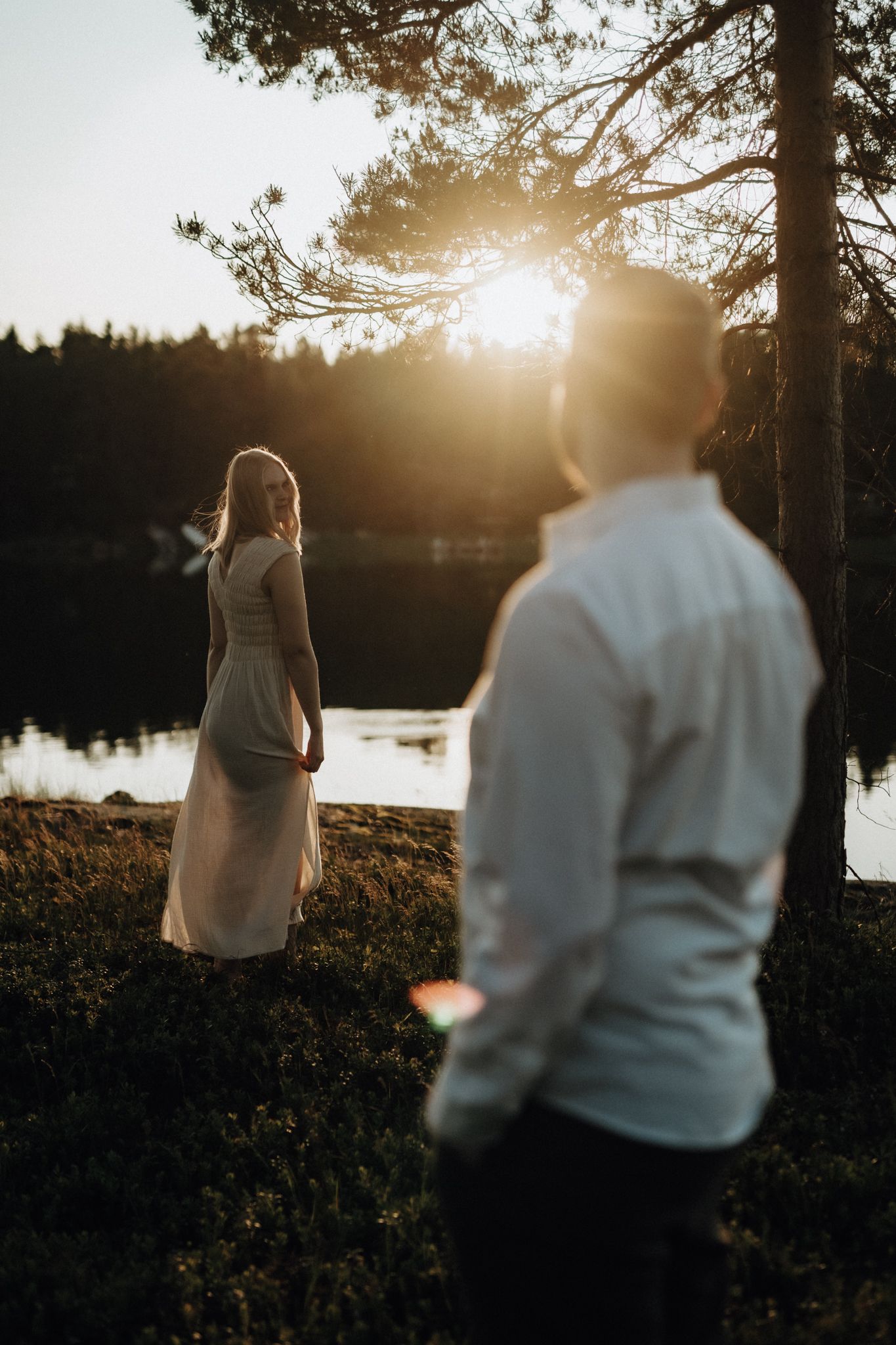 Woman in a beige dress standing in golden sunset light by a lake with man in foreground.