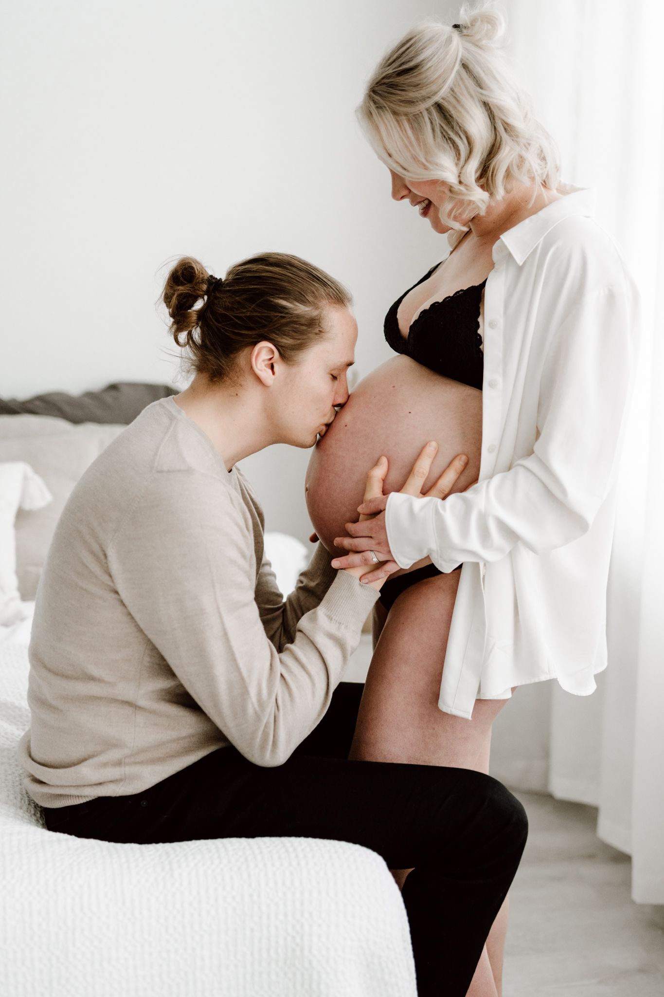 Man sits on bed kissing pregnant woman's bare belly while she smiles down at him lovingly.