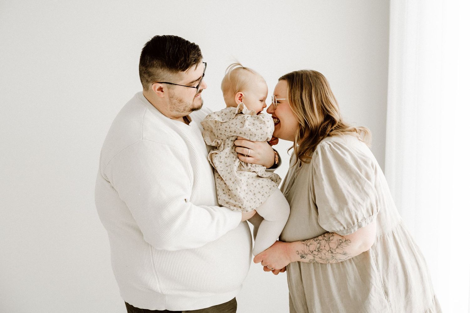 A couple in neutral tones share a tender moment with their baby girl in a bright white studio setting.