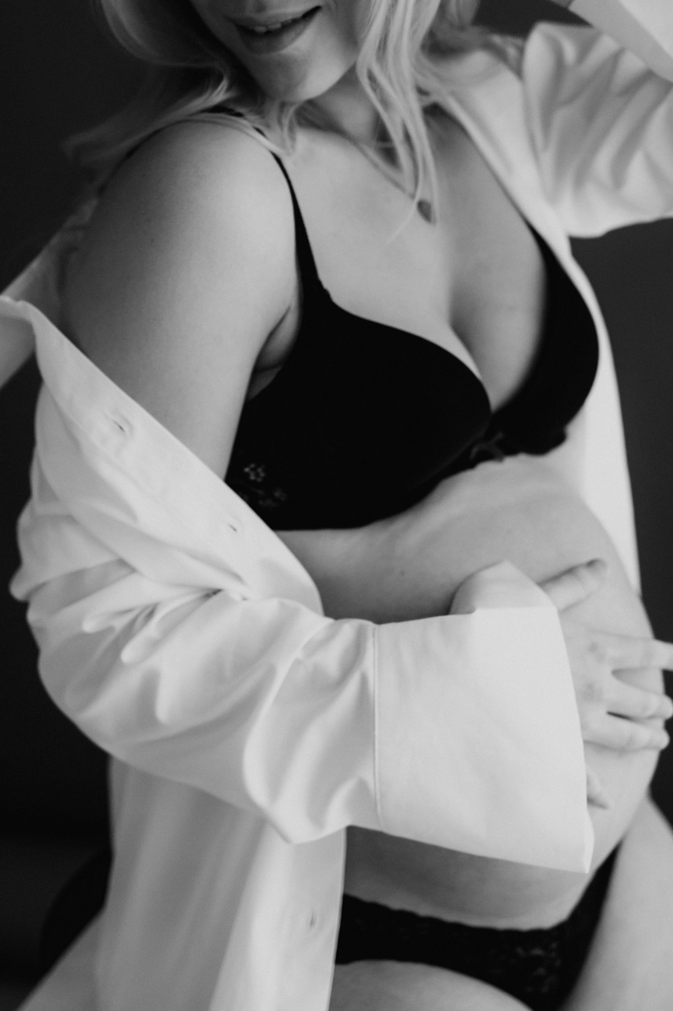 Black and white photo of pregnant woman in black bra and open white shirt posing for maternity boudoir.