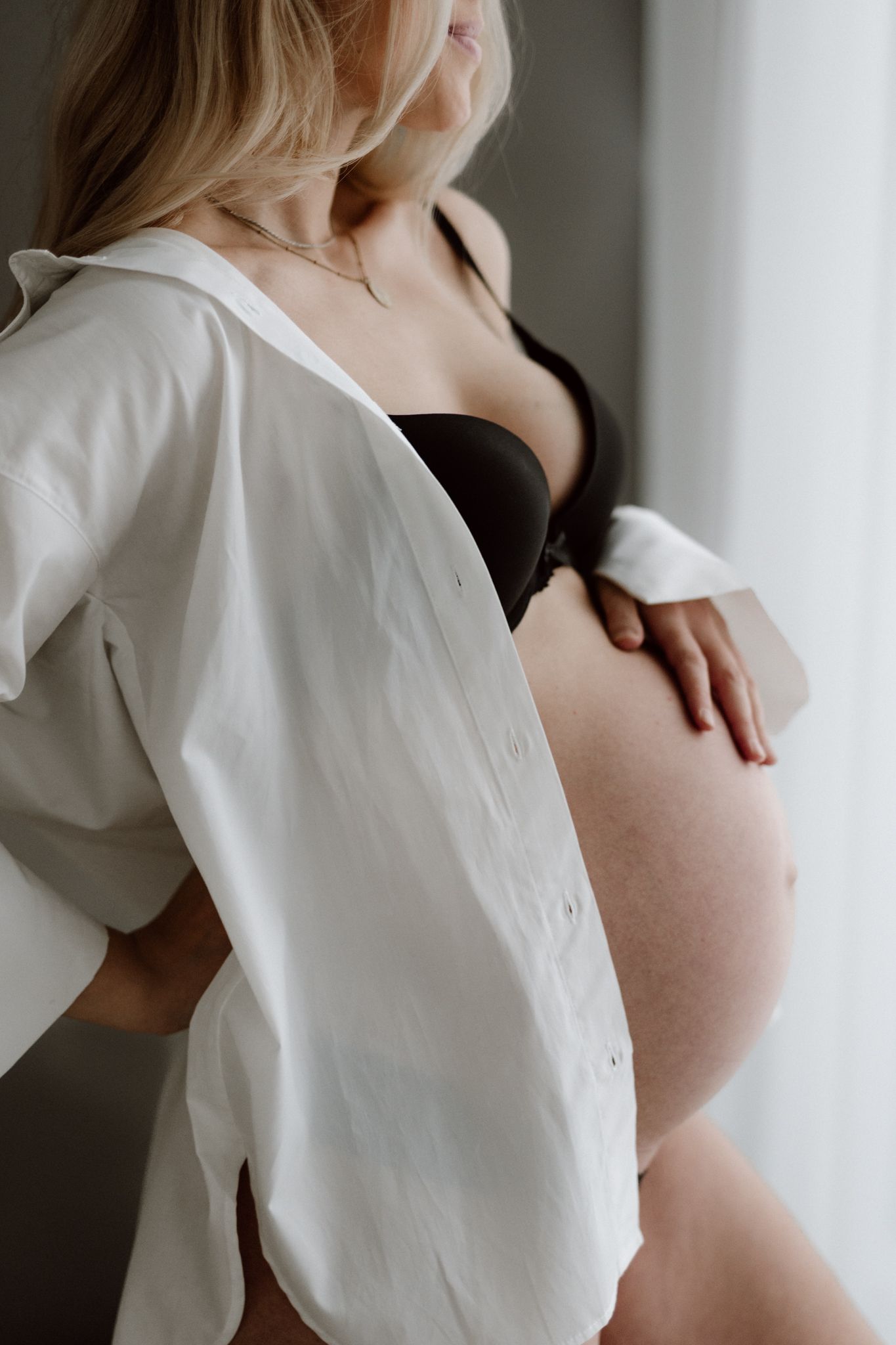Pregnant woman in open white shirt and black bra gently holding bare baby bump near bright window.