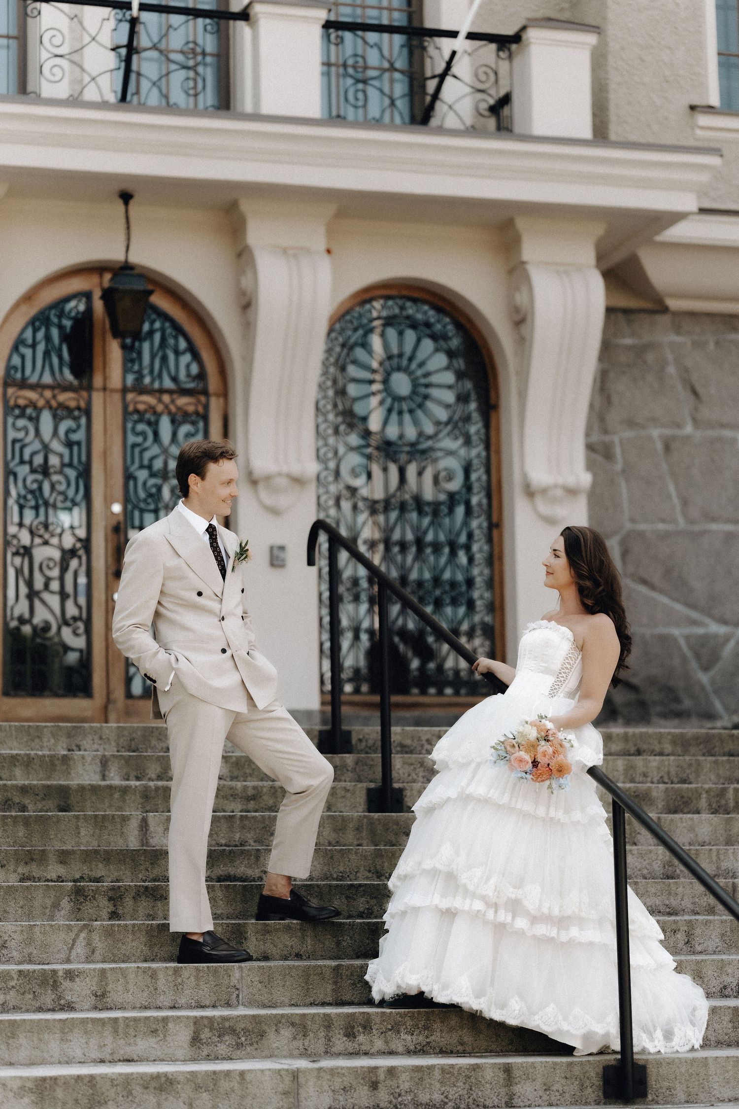 Bride and groom smiling at each other on stone steps outside an elegant building with arched windows.