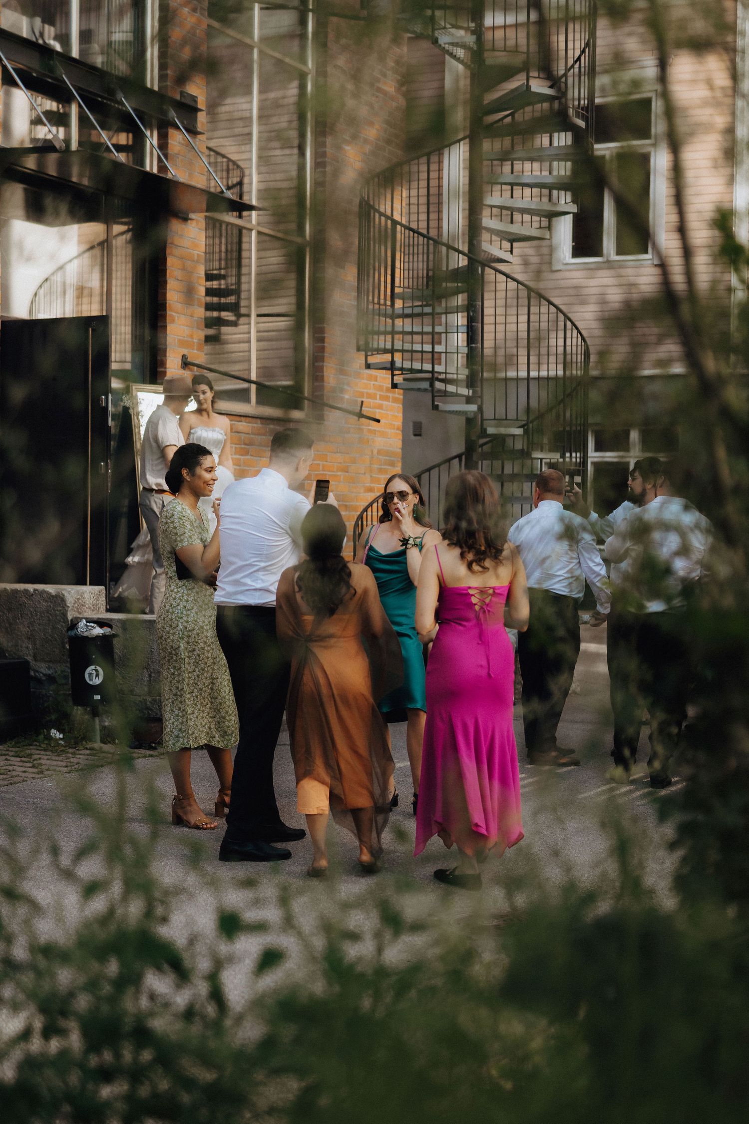 Elegantly dressed wedding guests mingle outdoors near a brick building with a spiral staircase.