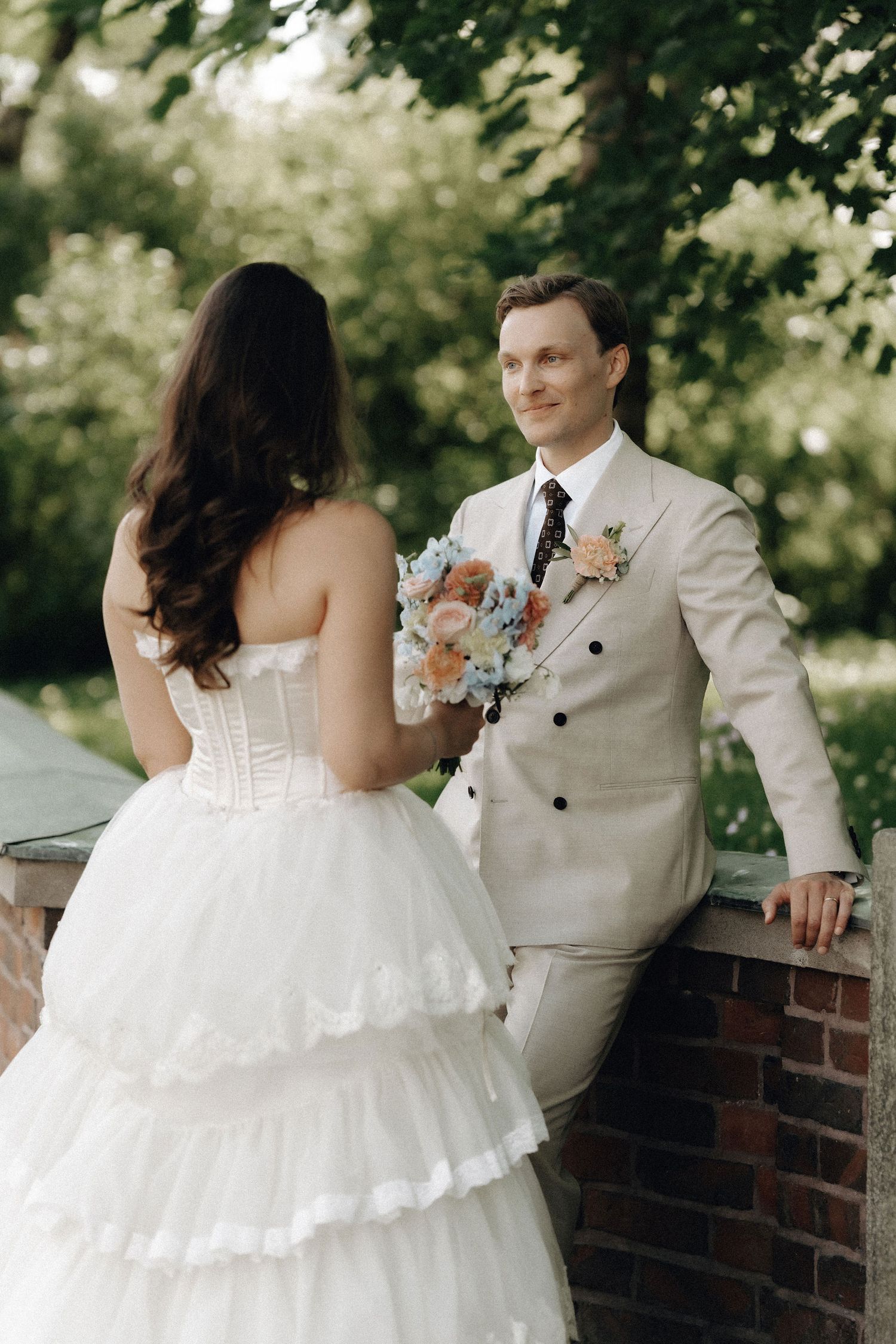 Groom in beige suit smiles at bride in layered white dress holding pastel bouquet in garden setting.