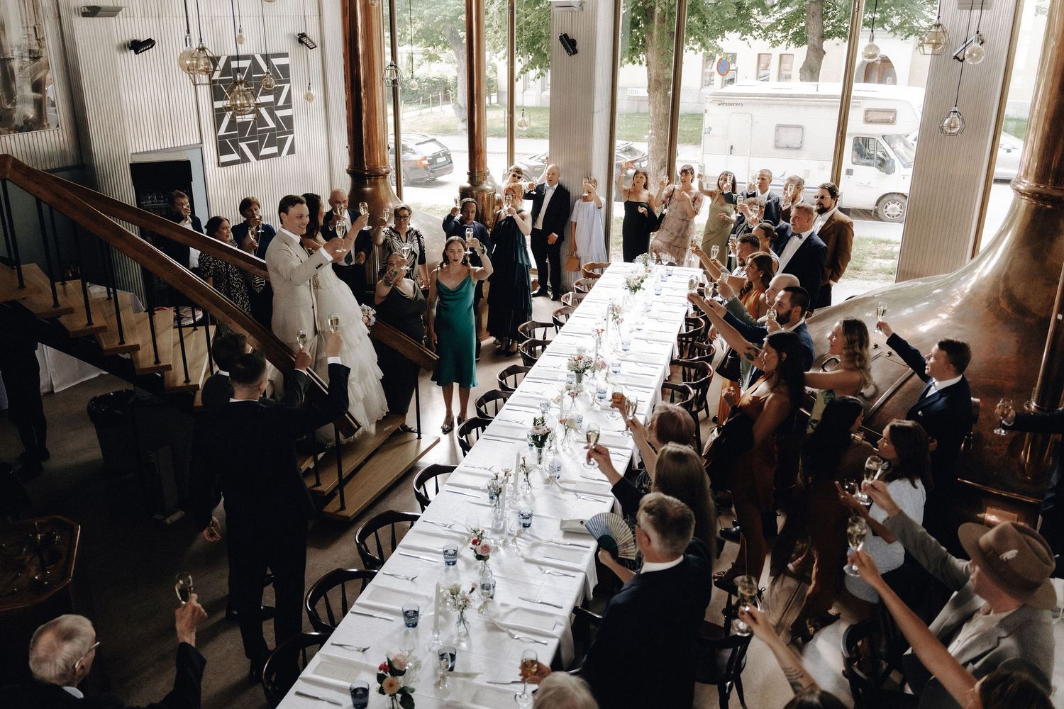 Aerial view of wedding reception with guests toasting at long elegantly decorated dinner table.