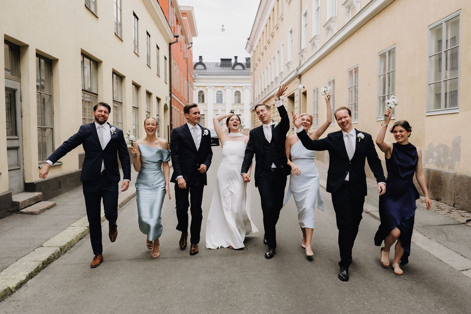 Iloinen hääseurue kävelemässä yhdessä mukulakivikadulla juhla-asuissa. Hääkuvaus Helsingissä Garden by Olossa.