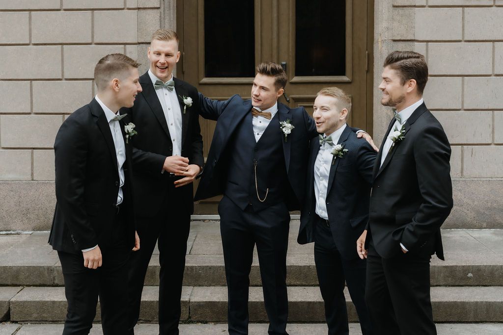 Five groomsmen in black suits with boutonnieres laugh outside a grand building.