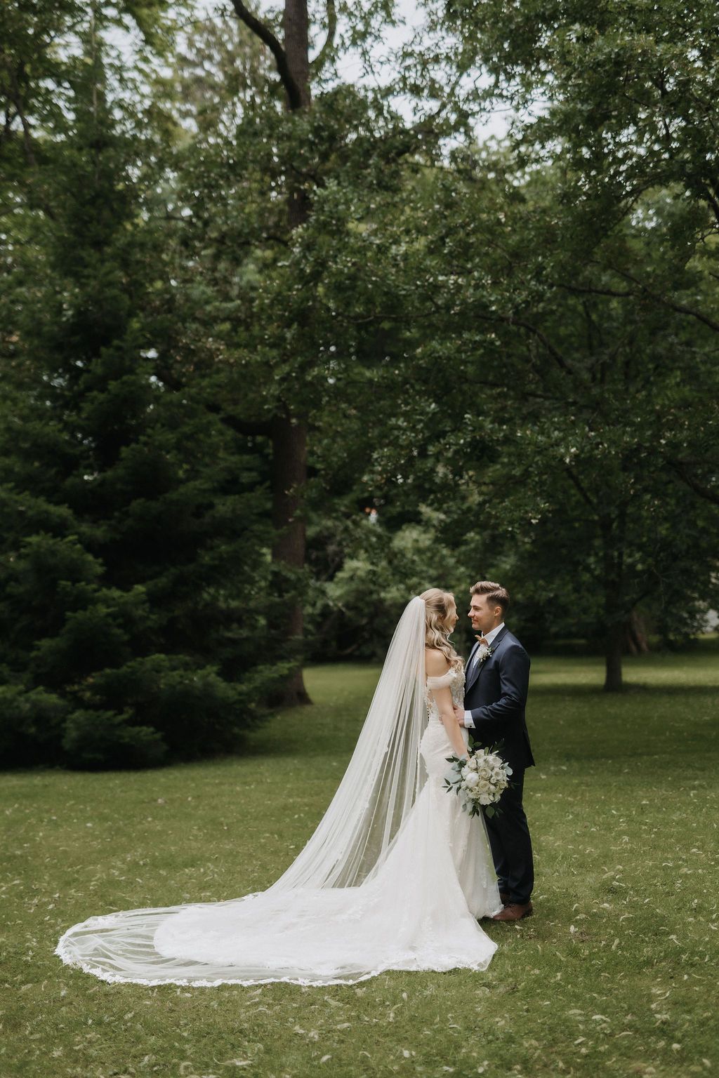 Bride and groom stand together on green lawn surrounded by lush trees during outdoor wedding portrait session.