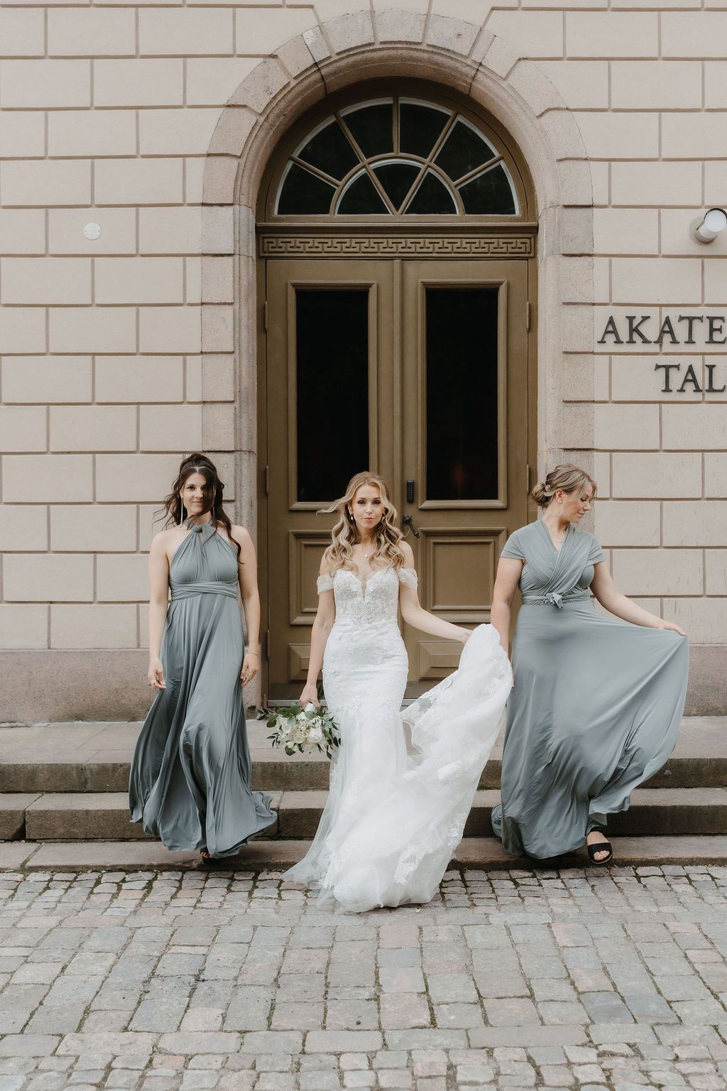 Bride in lace mermaid gown walks with two bridesmaids in sage dresses before Akatemia Talo entrance.
