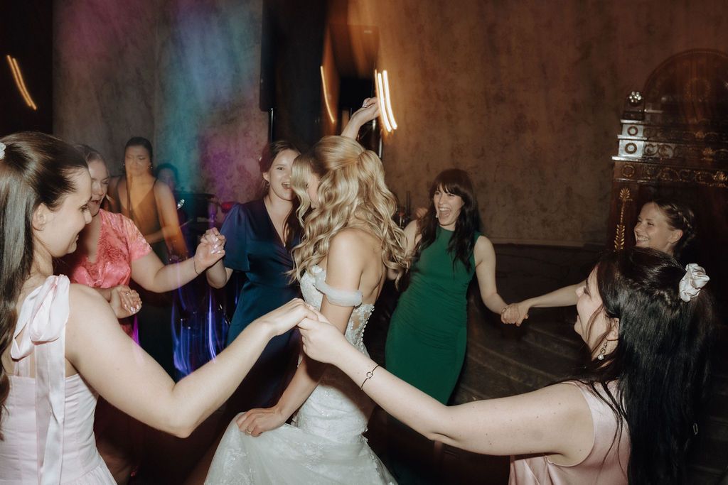 Bride surrounded by bridesmaids in circle dance at colorfully lit wedding reception.