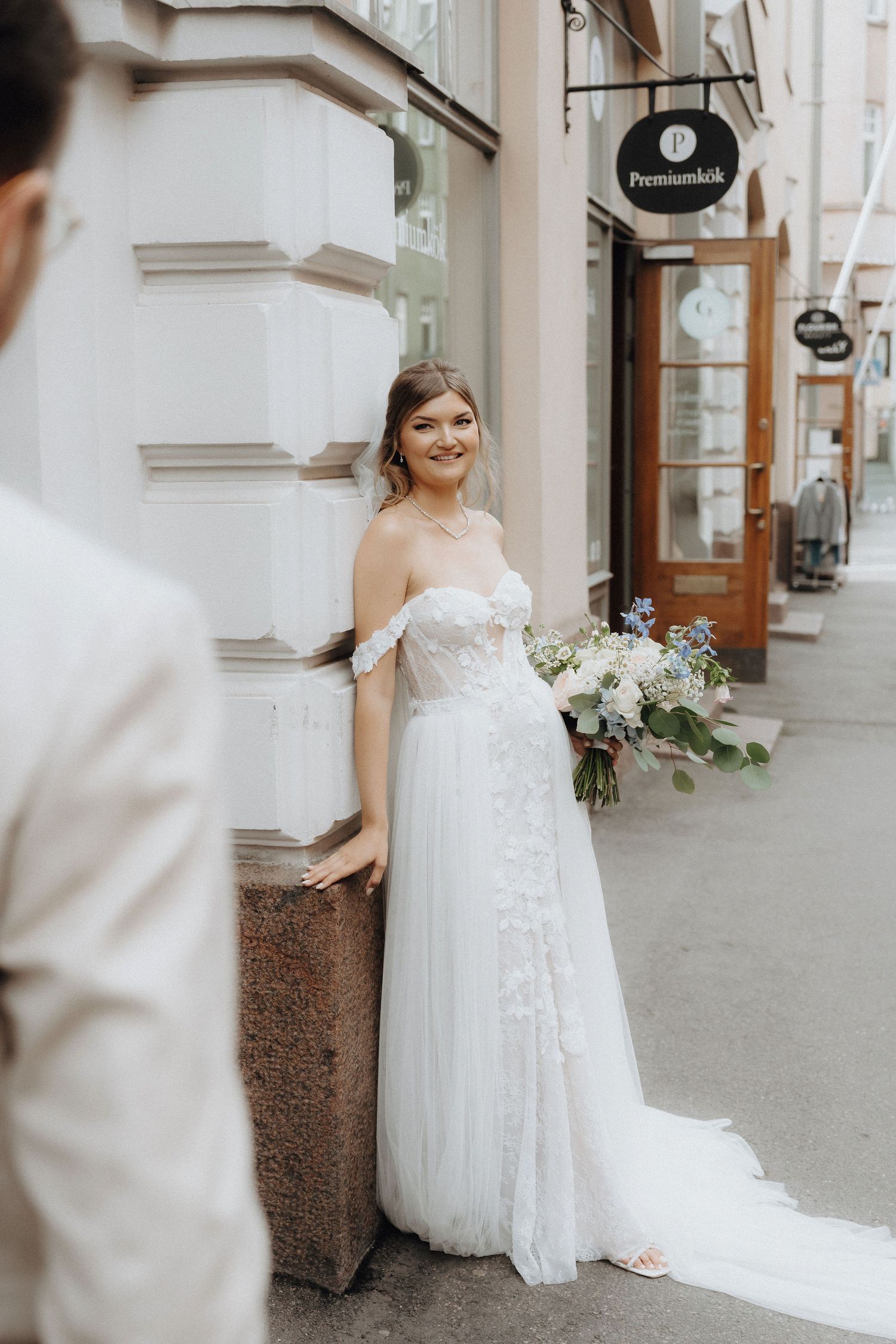 Smiling bride in off-shoulder lace wedding gown holding a blue and white bouquet on a city street.