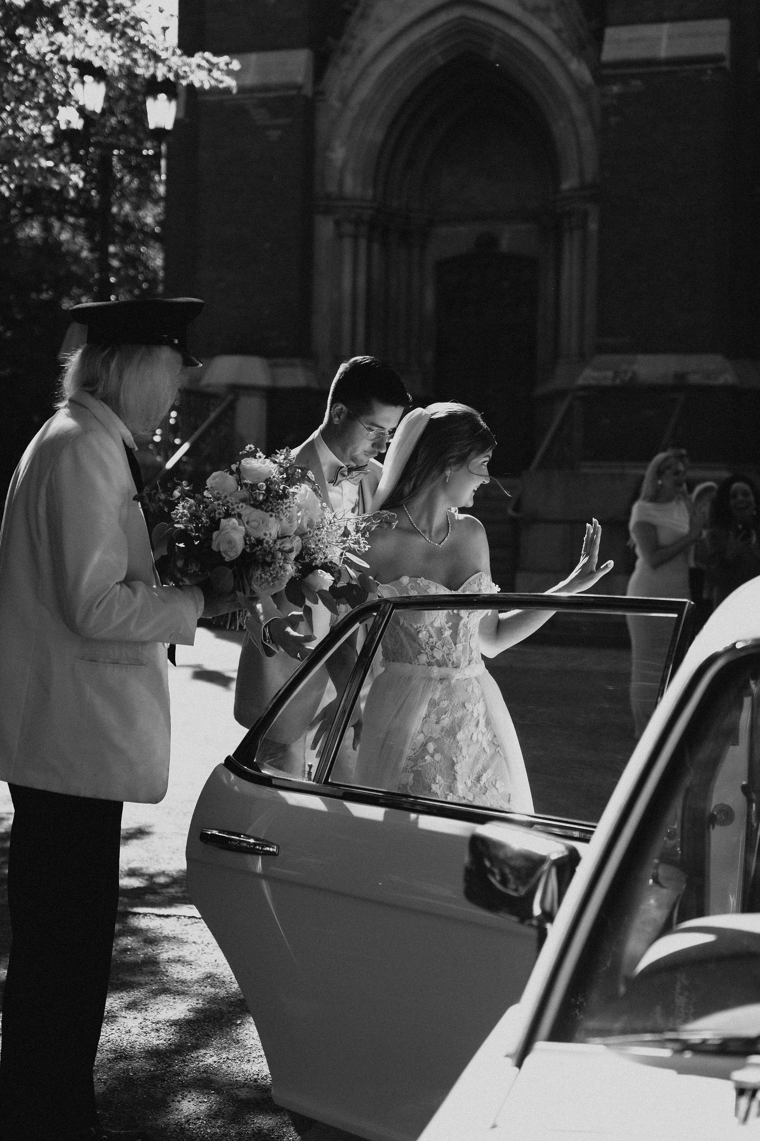 Black and white photo of bride and groom getting into a classic white car outside a church.