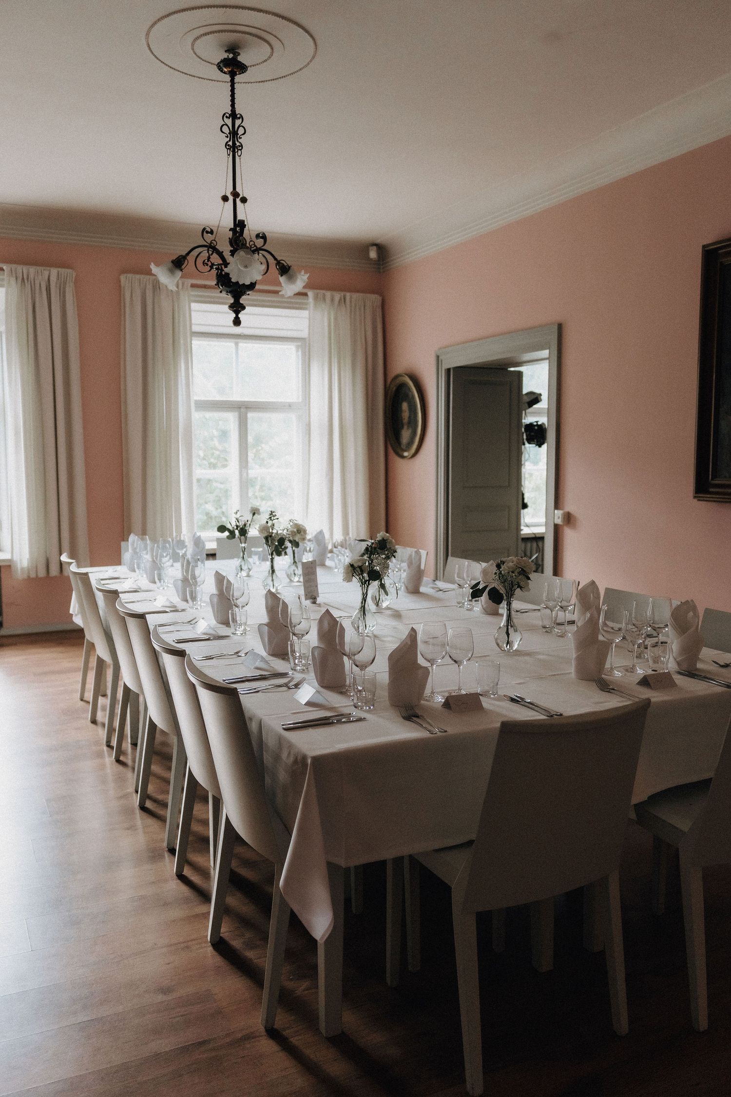 Long wedding banquet table with white tablecloths, white chairs, and floral arrangements in a pink-walled room.