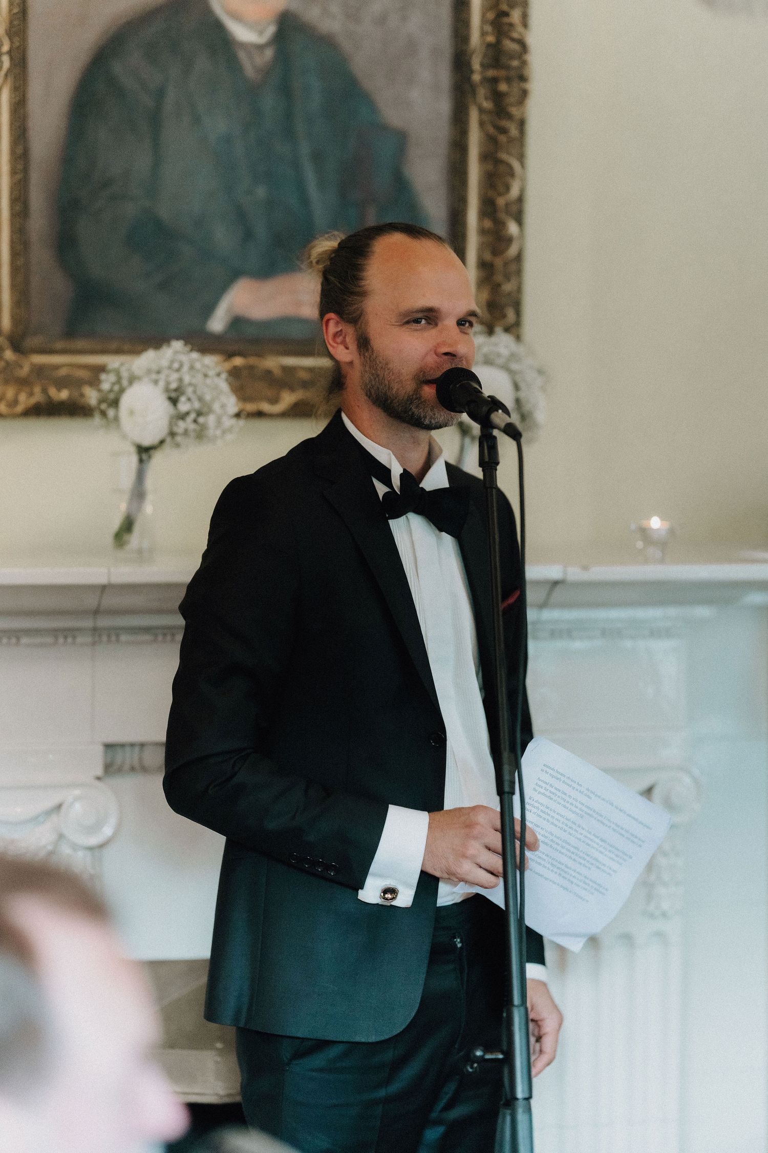 Elegant man in tuxedo giving a wedding speech at a microphone in a classic decorated room.