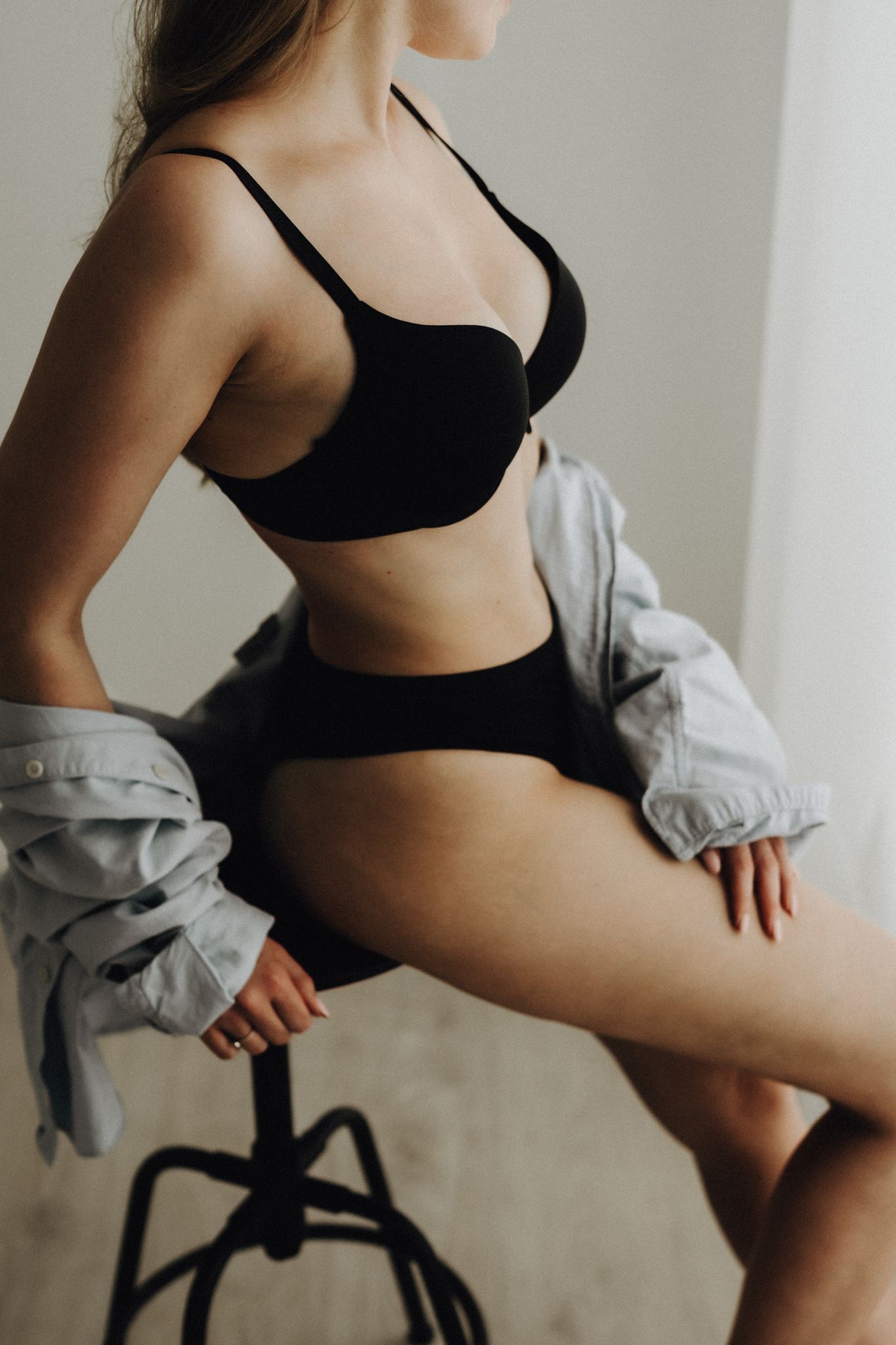Woman in black bra and underwear sitting on stool wearing open denim jacket.