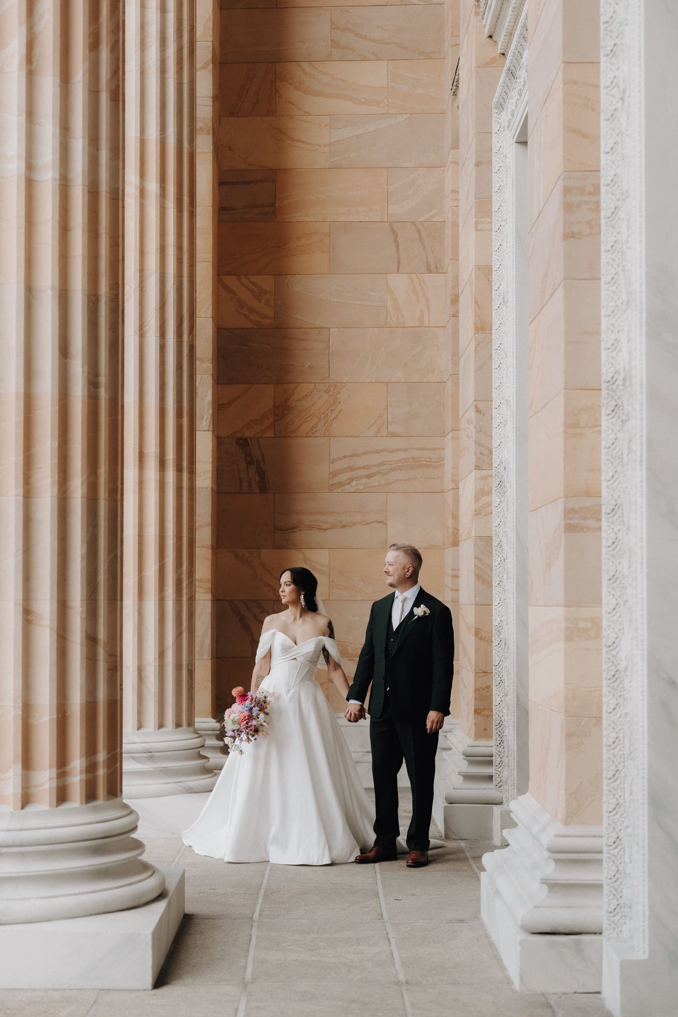 Bride in off-shoulder gown and groom in dark suit walking hand in hand between elegant marble columns.
