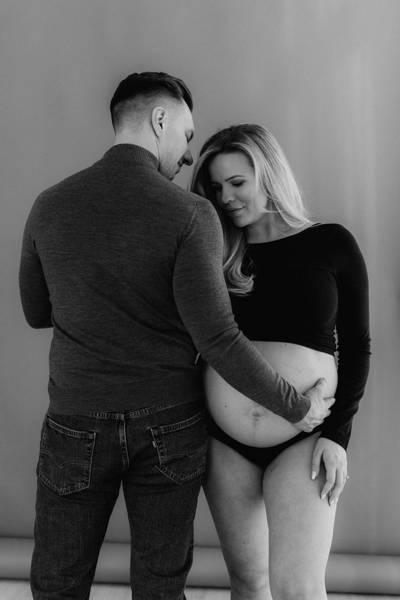 Black and white maternity photo of pregnant couple embracing, man touching baby bump in studio.