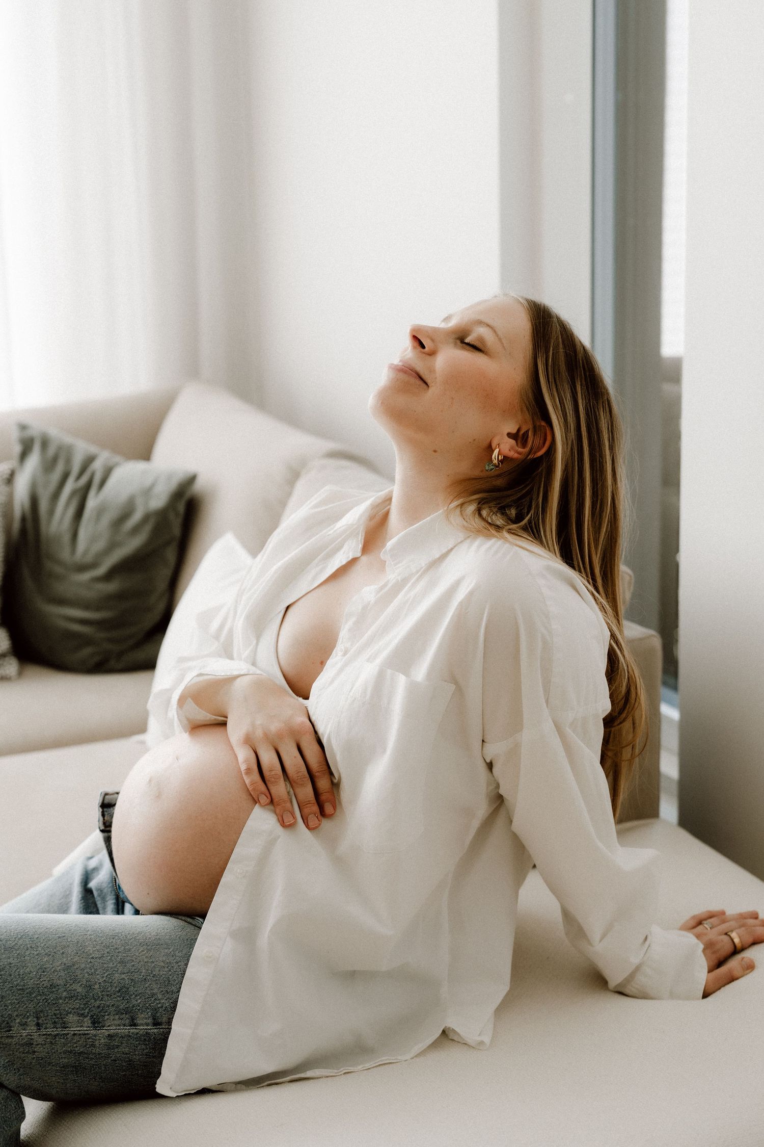 Pregnant woman in open white shirt sitting by window with eyes closed in peaceful moment.