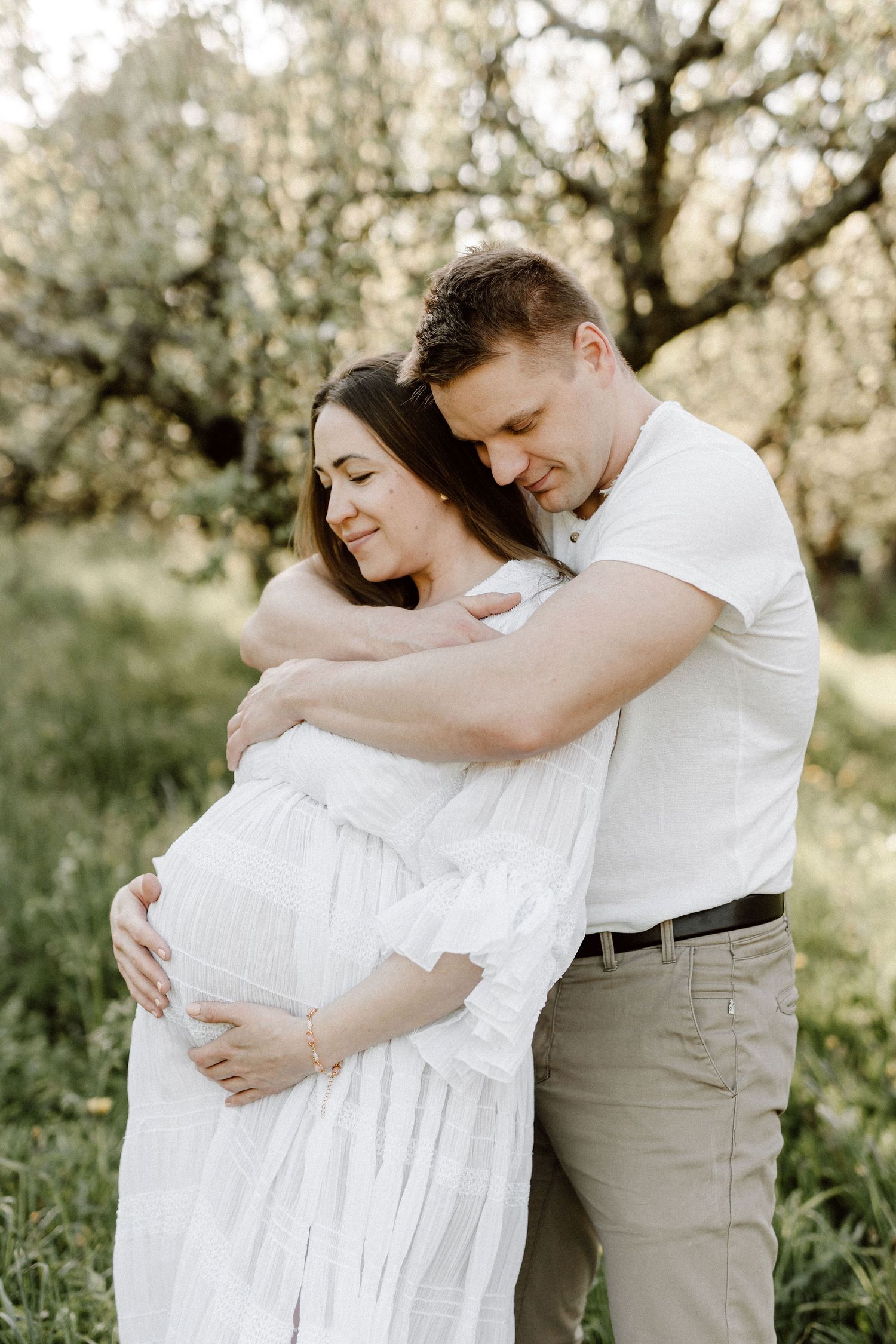 Man embracing pregnant woman from behind, both smiling softly in outdoor maternity photo.