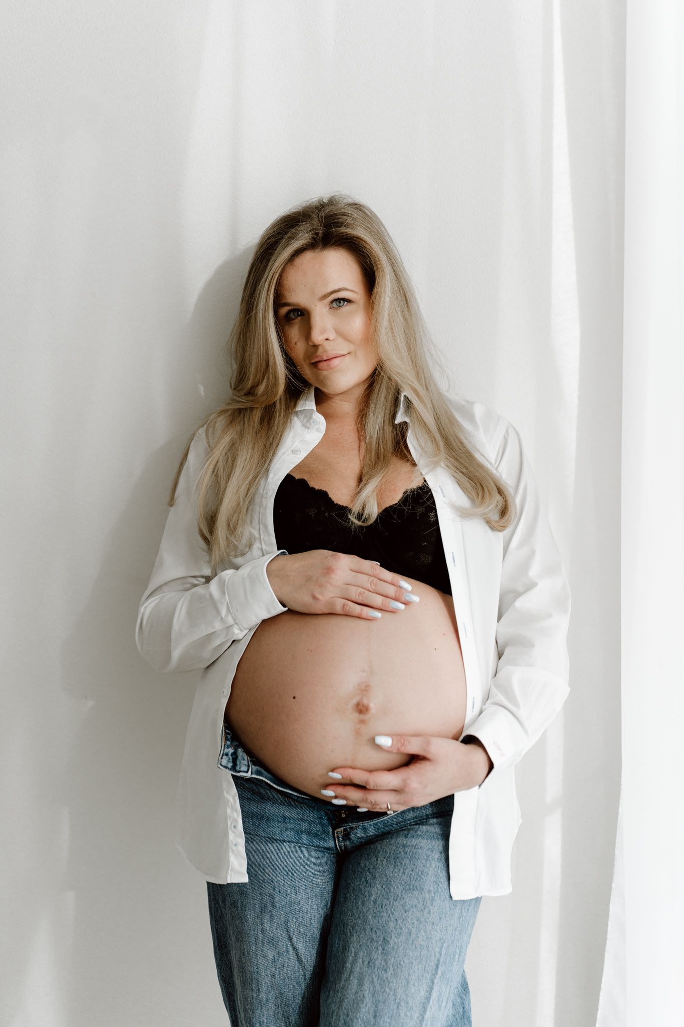 Blonde pregnant woman in black bra and white shirt cradling bare baby bump indoors.
