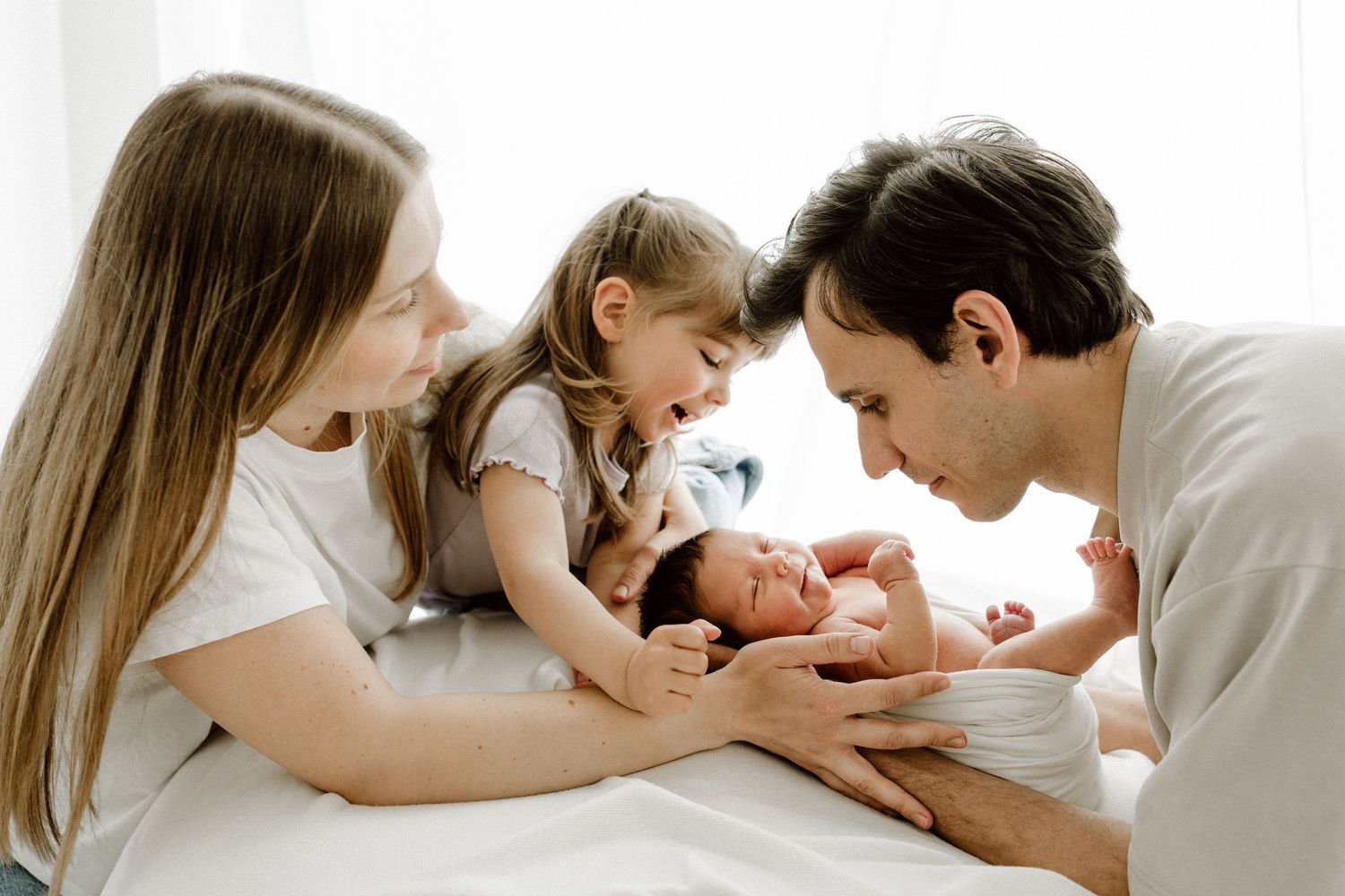 Happy family with mother, father, young daughter, and newborn baby on white bed.
