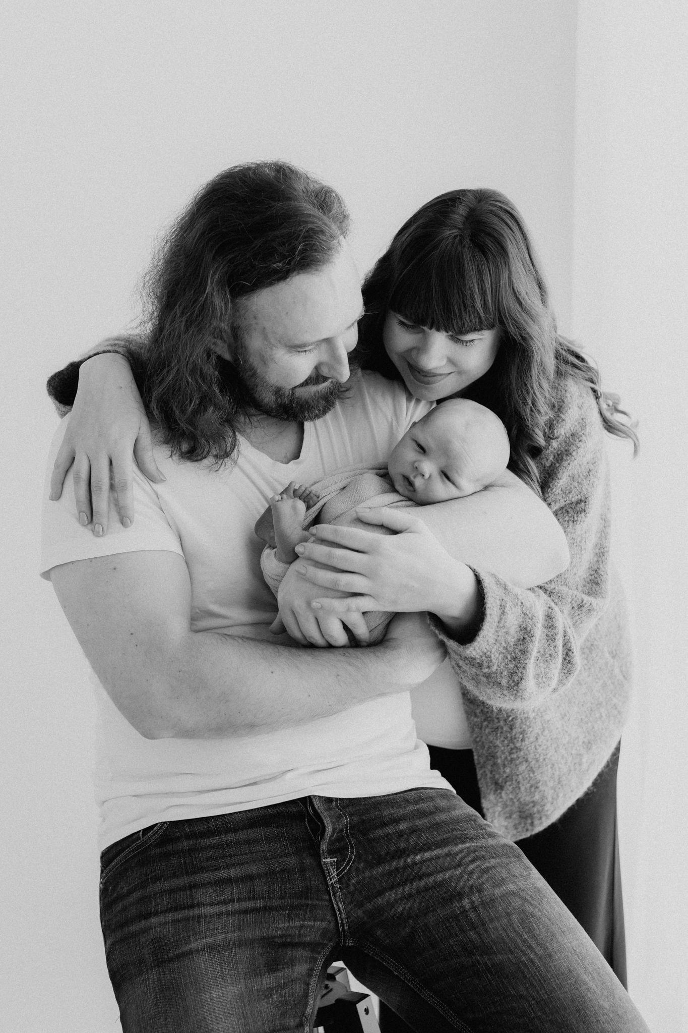 Black and white portrait of bearded father and brunette mother tenderly holding their newborn baby.