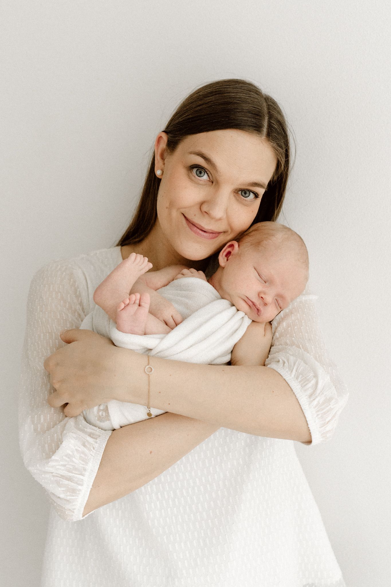 Smiling brunette mother in white dress holding swaddled sleeping newborn baby close to her chest.