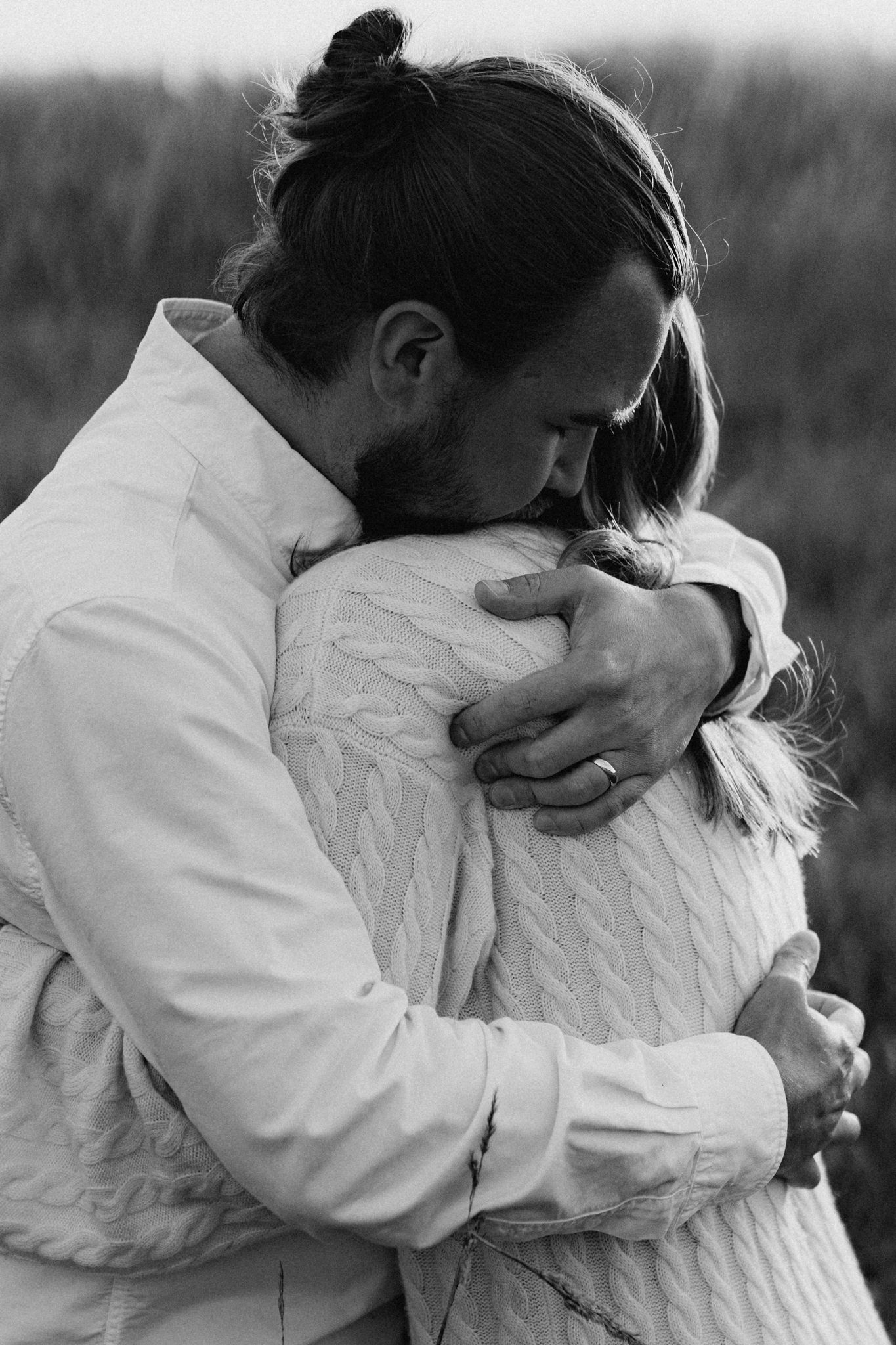 Black and white close-up of a bearded man with bun embracing a woman tenderly in a grassy field.
