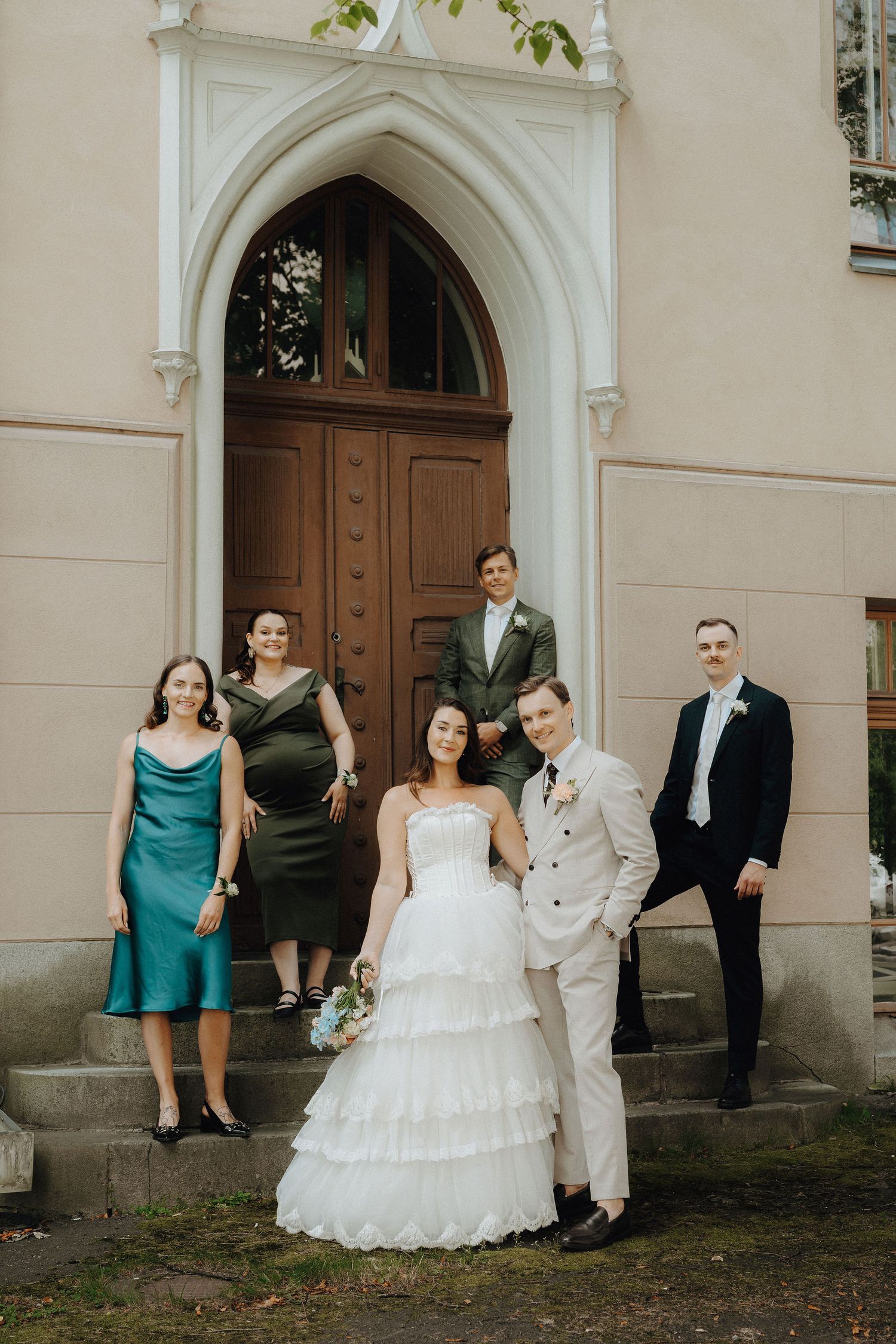 Wedding party of five posing on church steps, bride in tiered white gown with groom in beige suit.