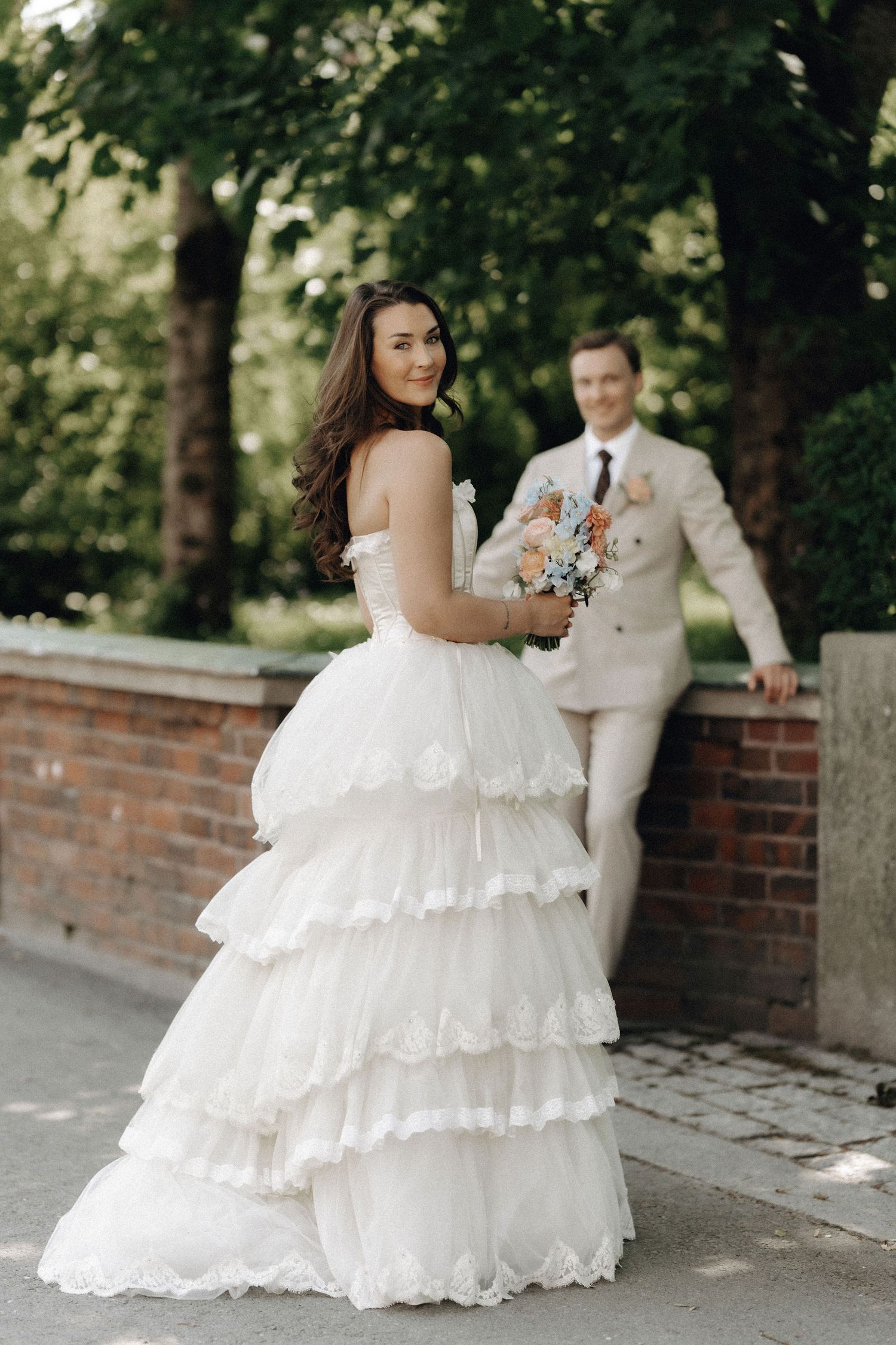 Bride in elegant tiered white gown holds pastel bouquet while groom in beige suit stands behind her.