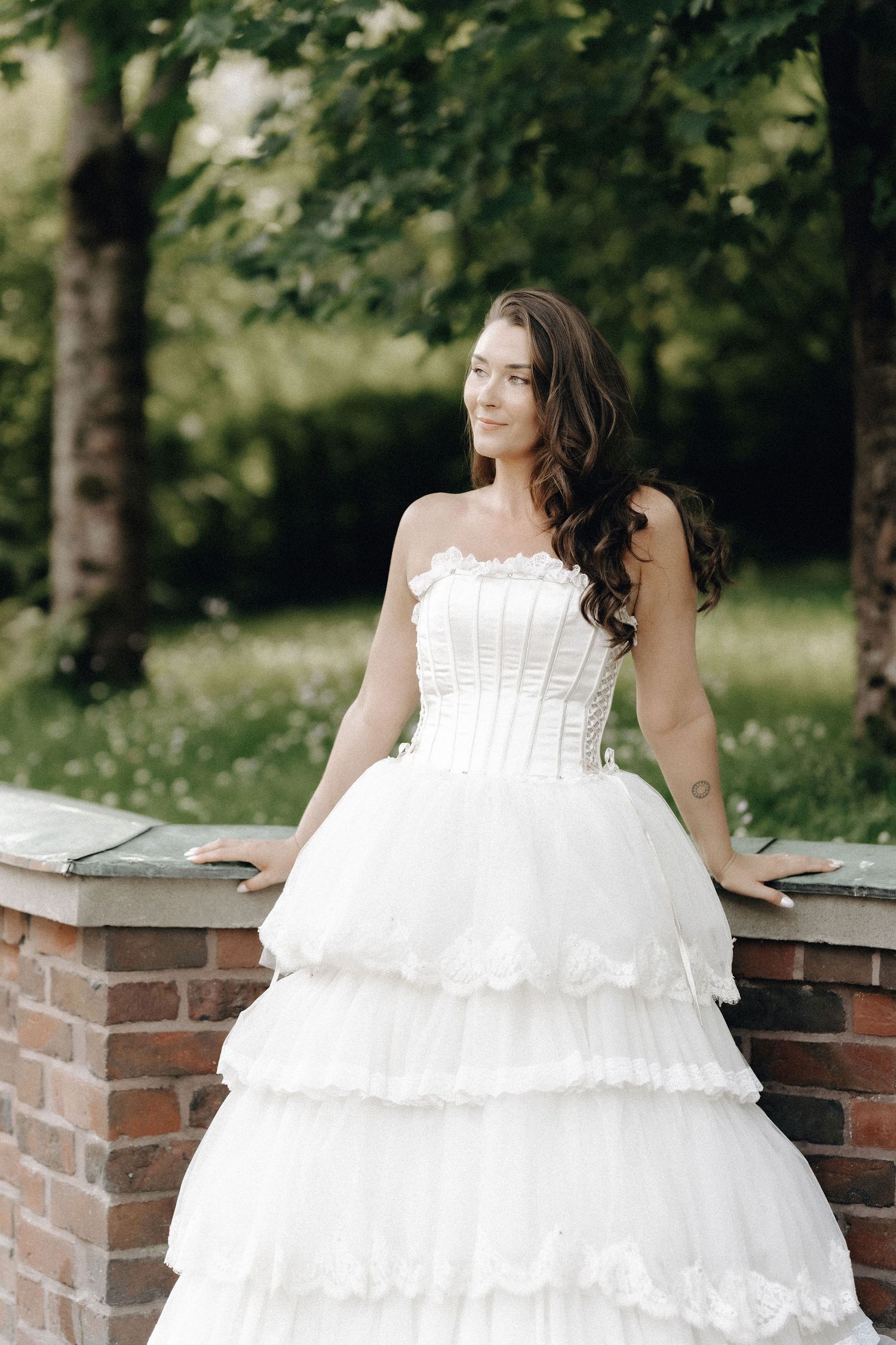 Beautiful bride in strapless tiered white wedding gown posing outdoors in green garden setting.